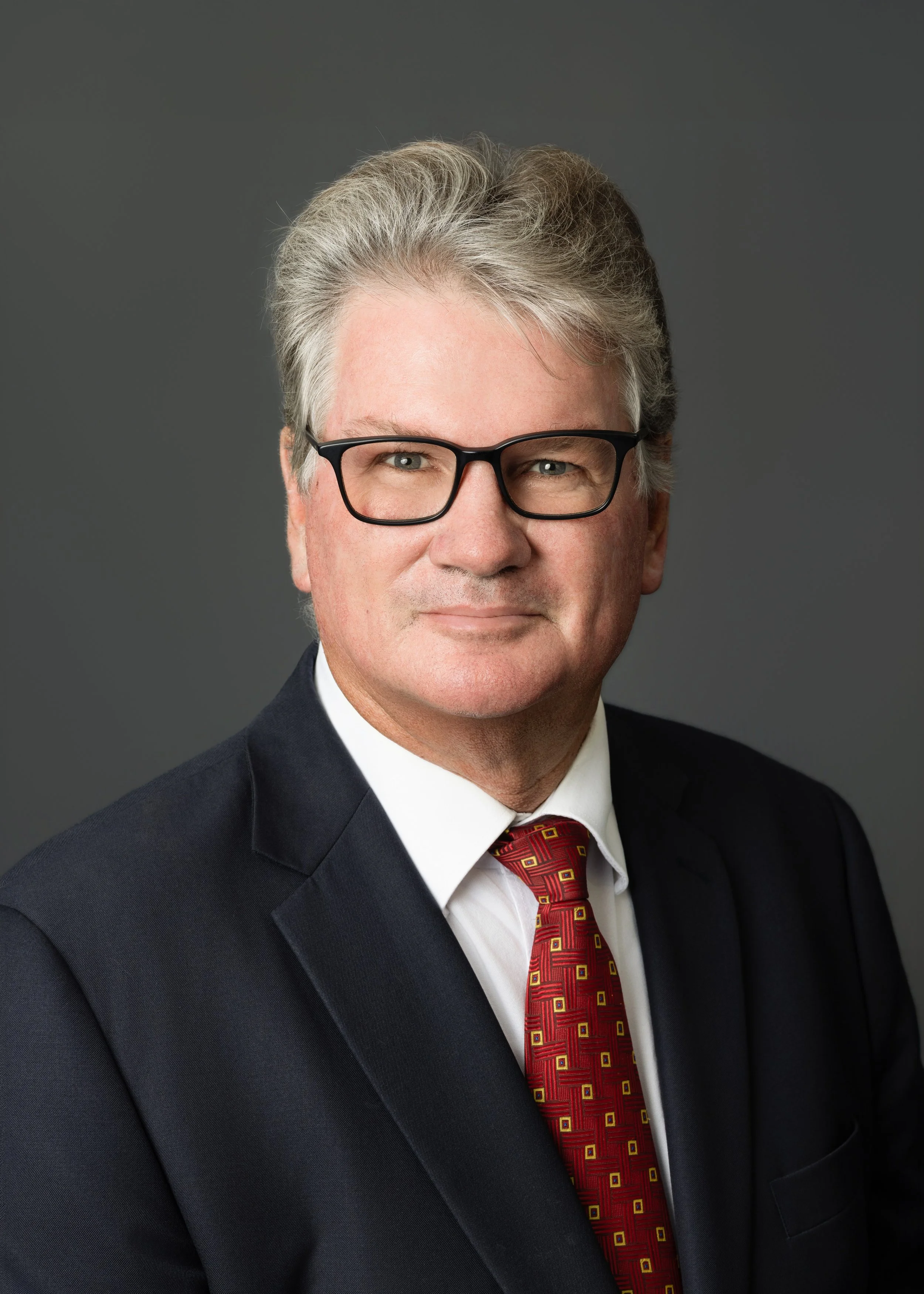 A middle-aged man wearing glasses, a dark suit, a white shirt, and a colorful patterned tie, standing against a plain background.