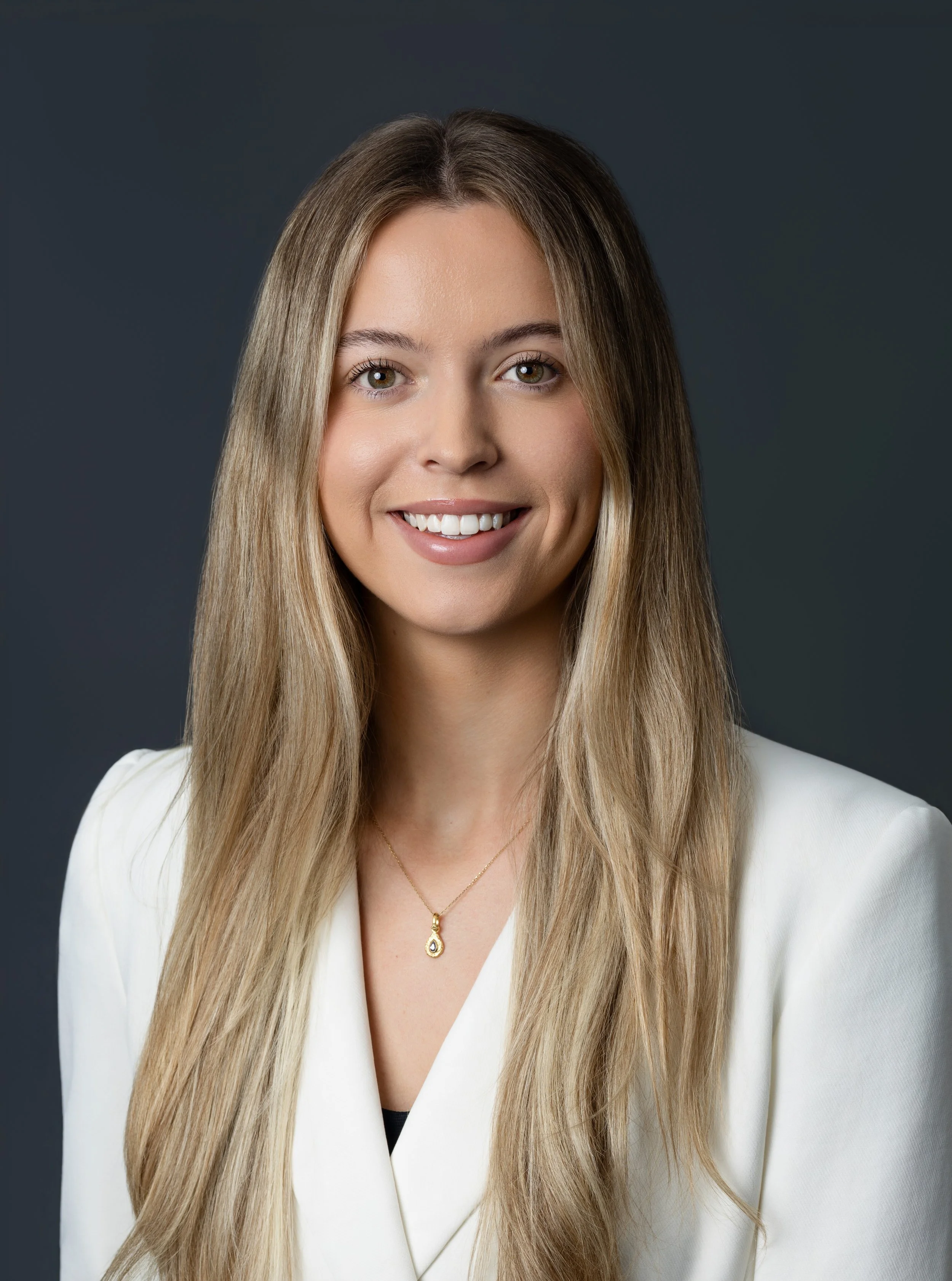 A young woman with long brown hair, wearing a black blazer and a white scarf, stands with crossed arms against a plain white background, smiling confidently.