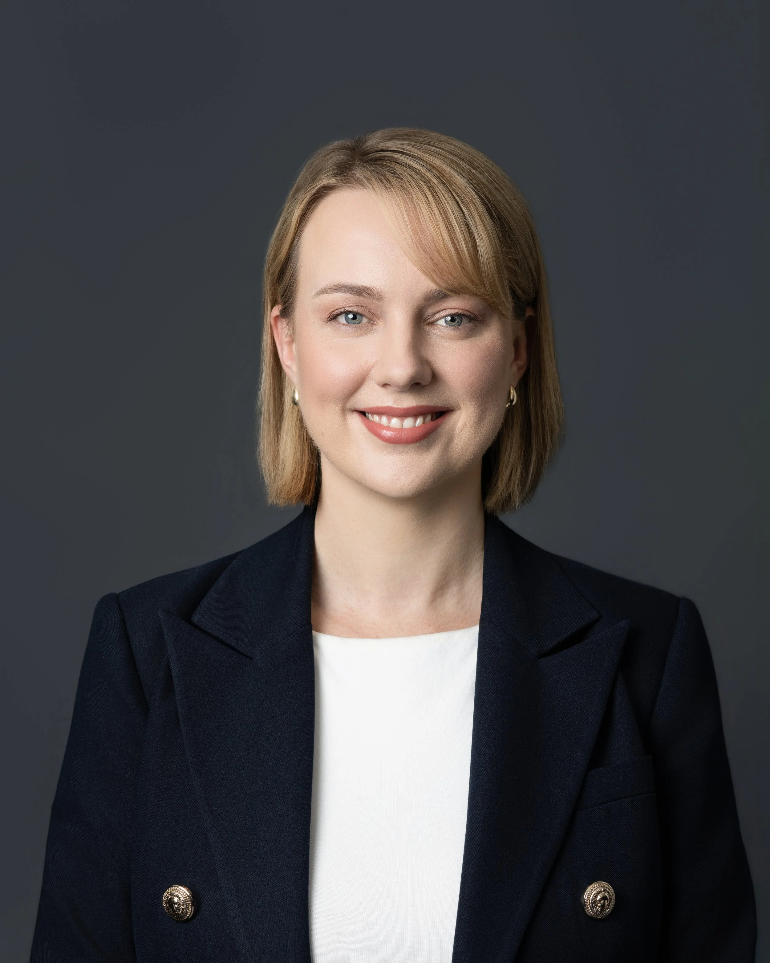 Black and white photo of a woman with shoulder-length hair, smiling, in an office setting with a plant and chair in the background.