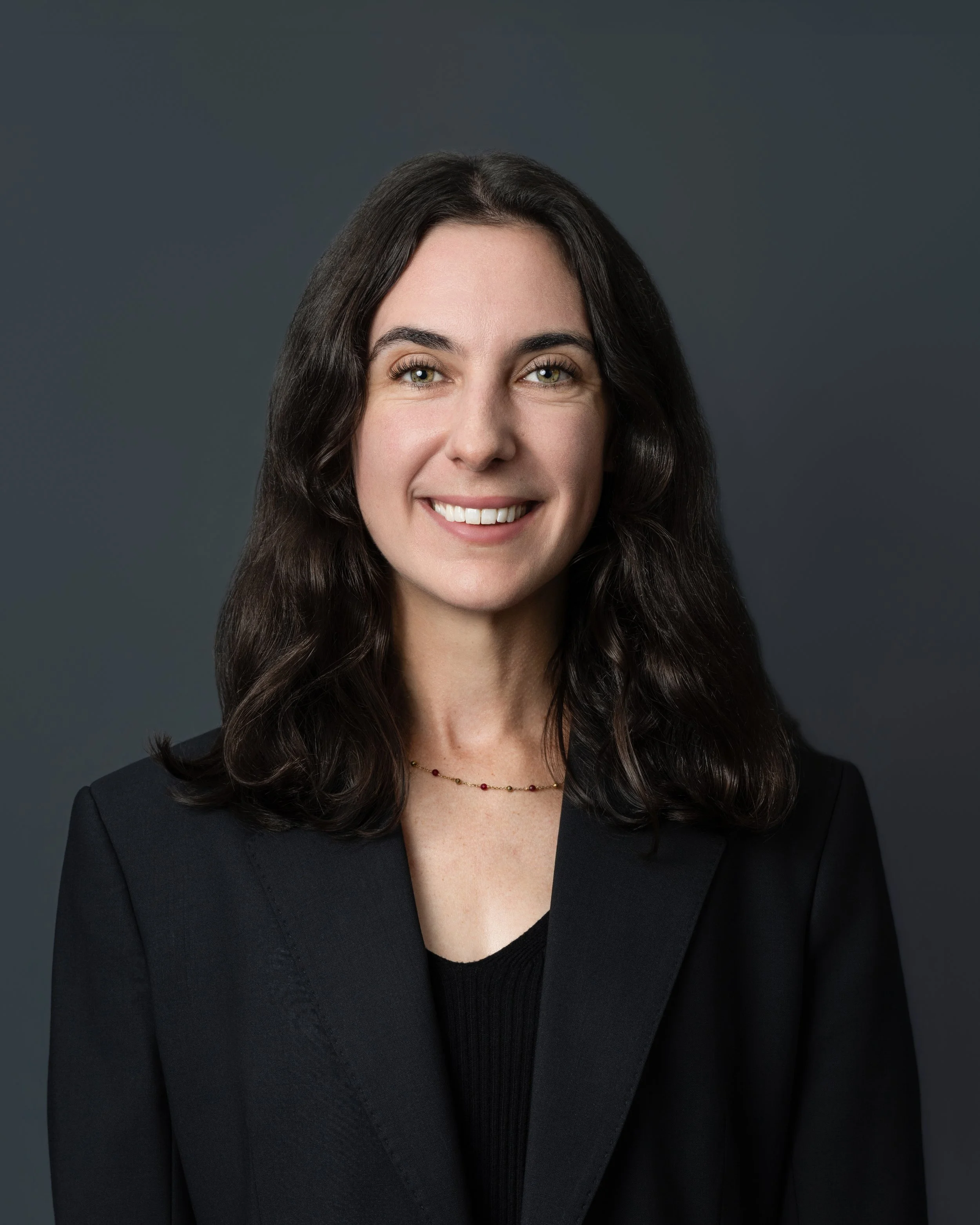 Smiling woman with dark, shoulder-length, wavy hair, wearing a white button-up shirt and hoop earrings, standing against a neutral background.