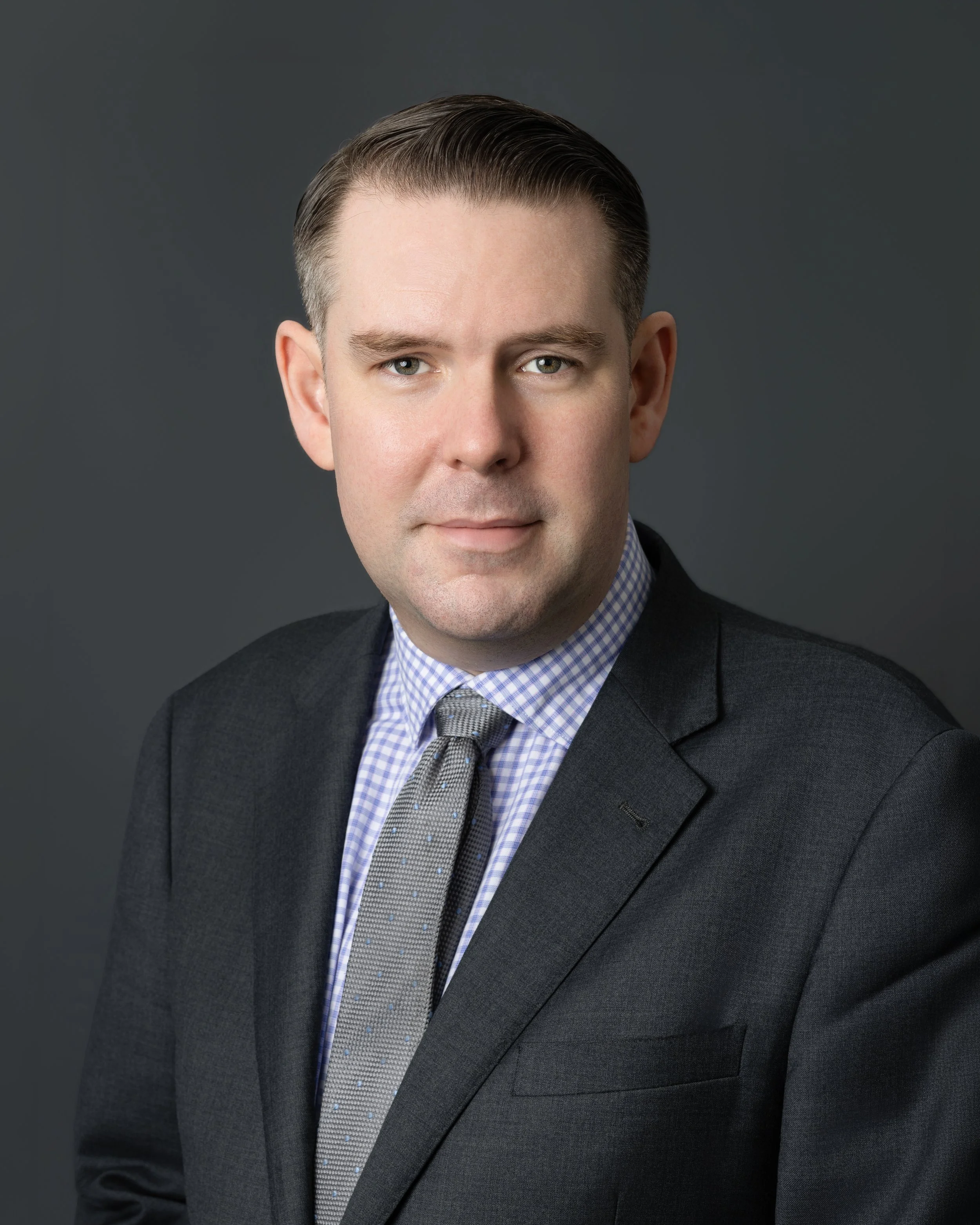A man in a dark suit with a checkered shirt and tie, smiling at the camera.