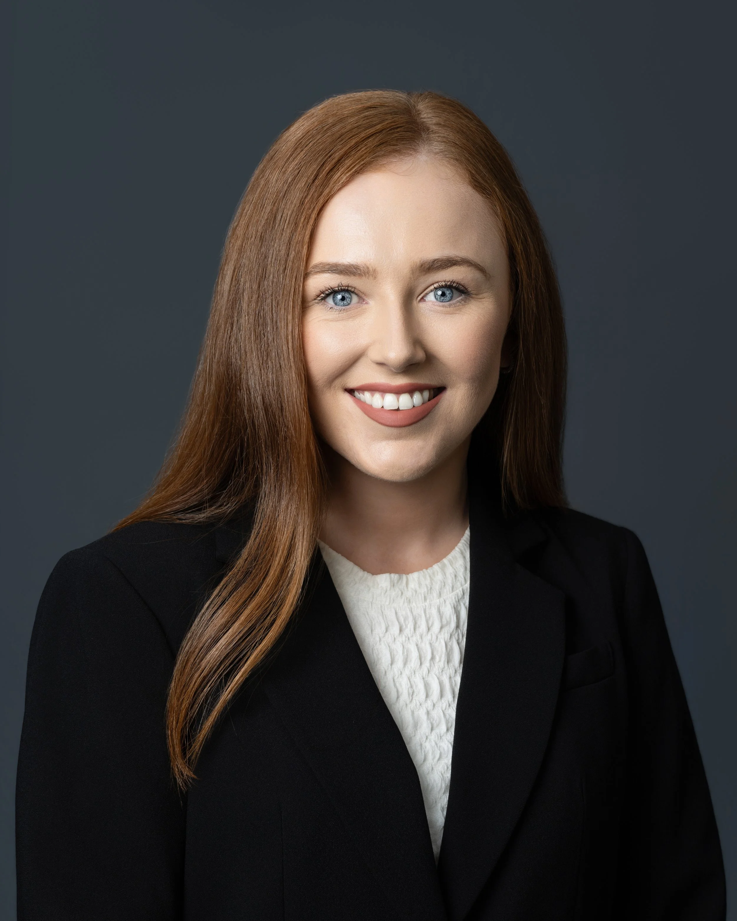 A young woman with long red hair smiling, wearing a black and white patterned blouse and black pants.