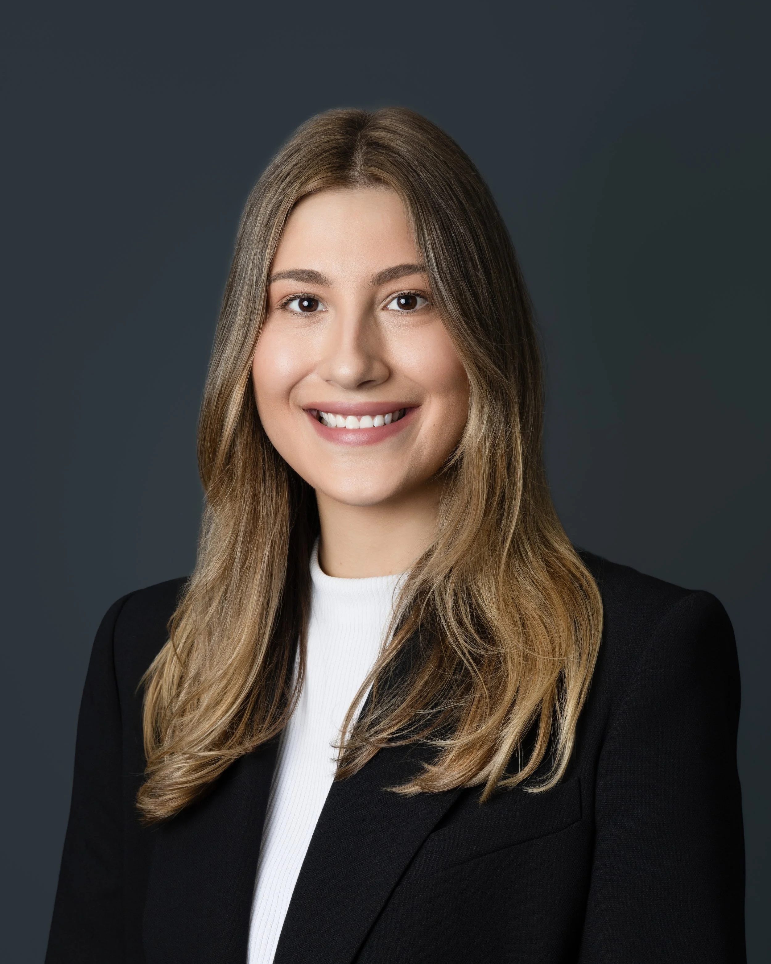 A young woman with long wavy blonde hair, smiling, wearing a black button-up shirt and a delicate necklace.