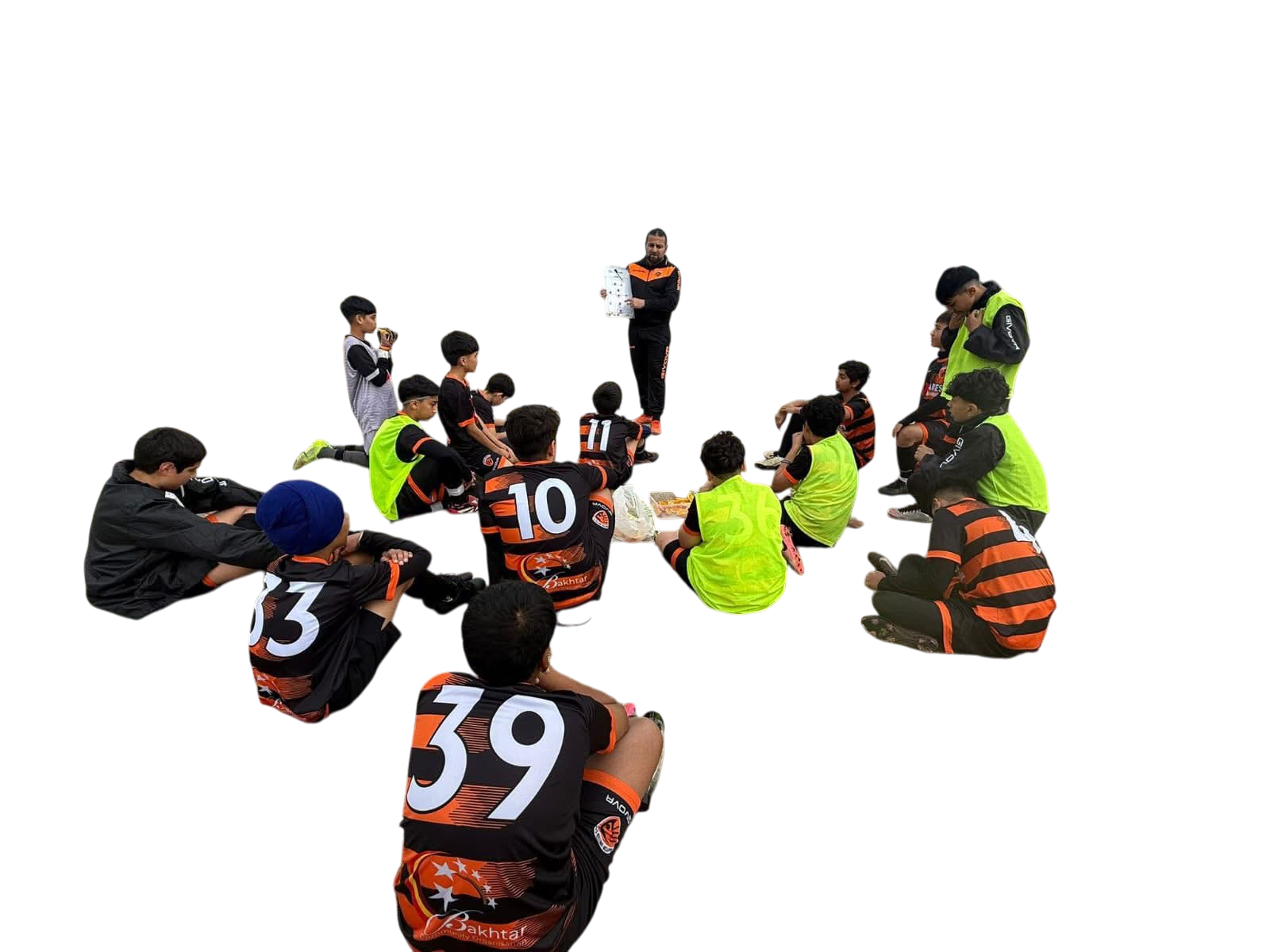 A group of young soccer players sitting on the ground in a semi-circle around a coach, during a team meeting or warm-up session. Players are wearing black and orange uniforms, some with bright yellow vests, and a few are wearing caps or headgear. The coach is standing with a clipboard, talking to them in an outdoor setting.