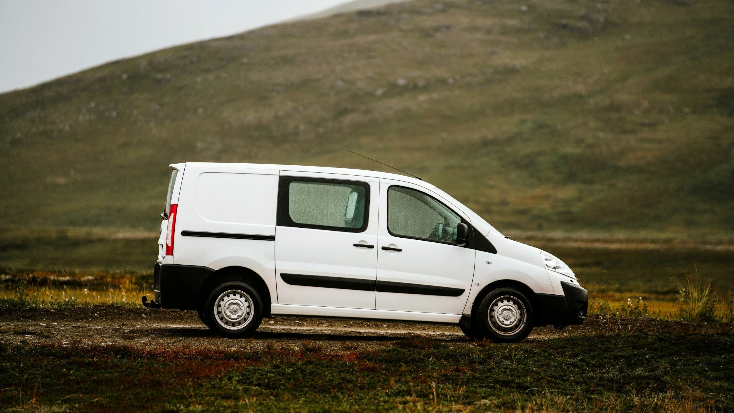 A white cargo van parked on a dirt road in a grassy, hilly landscape with overcast skies.