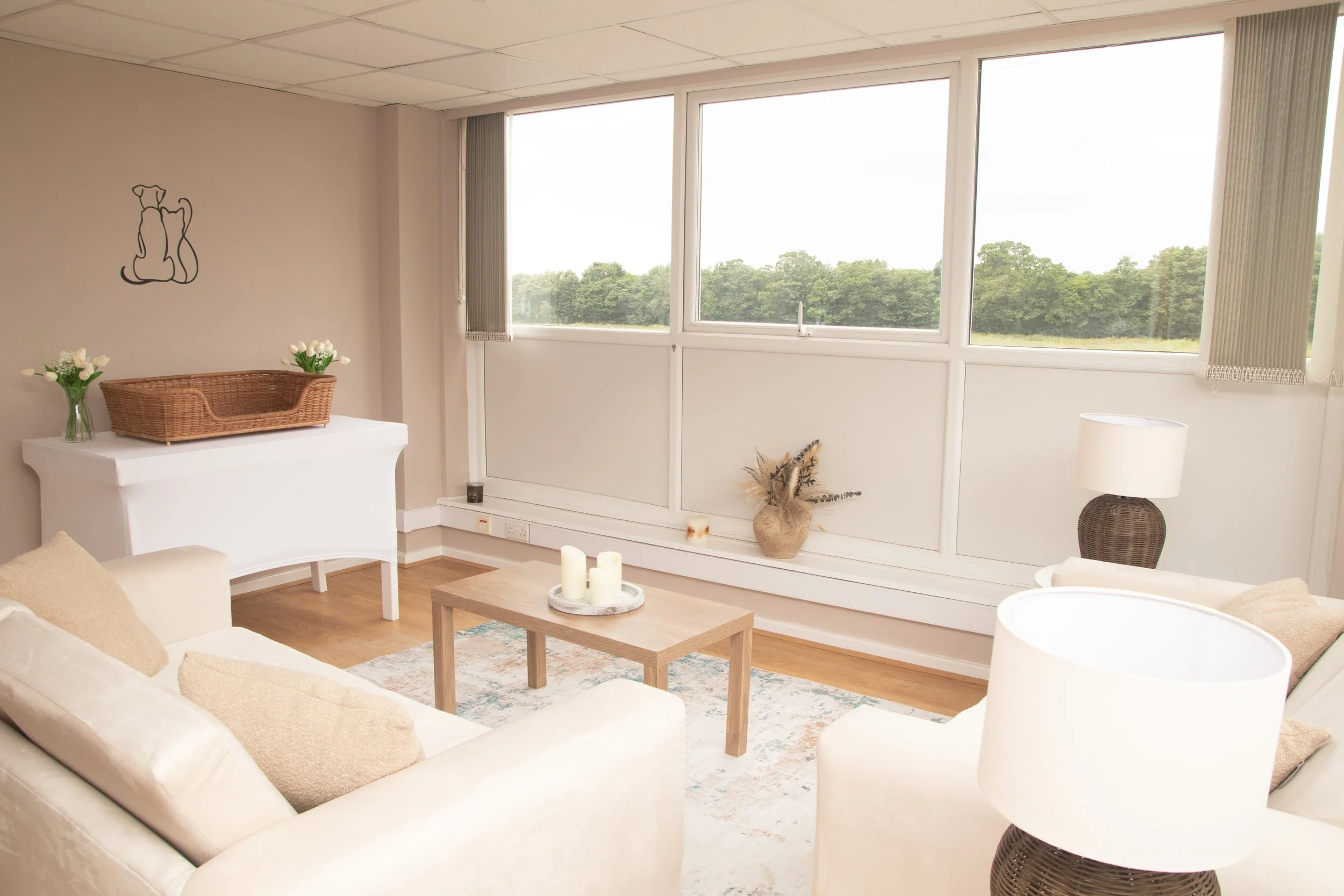 A living room with white sofas, beige cushions, a wooden coffee table with candles, a white piano with a wicker basket and flowers on top, a lamp, and large windows showing greenery outside.