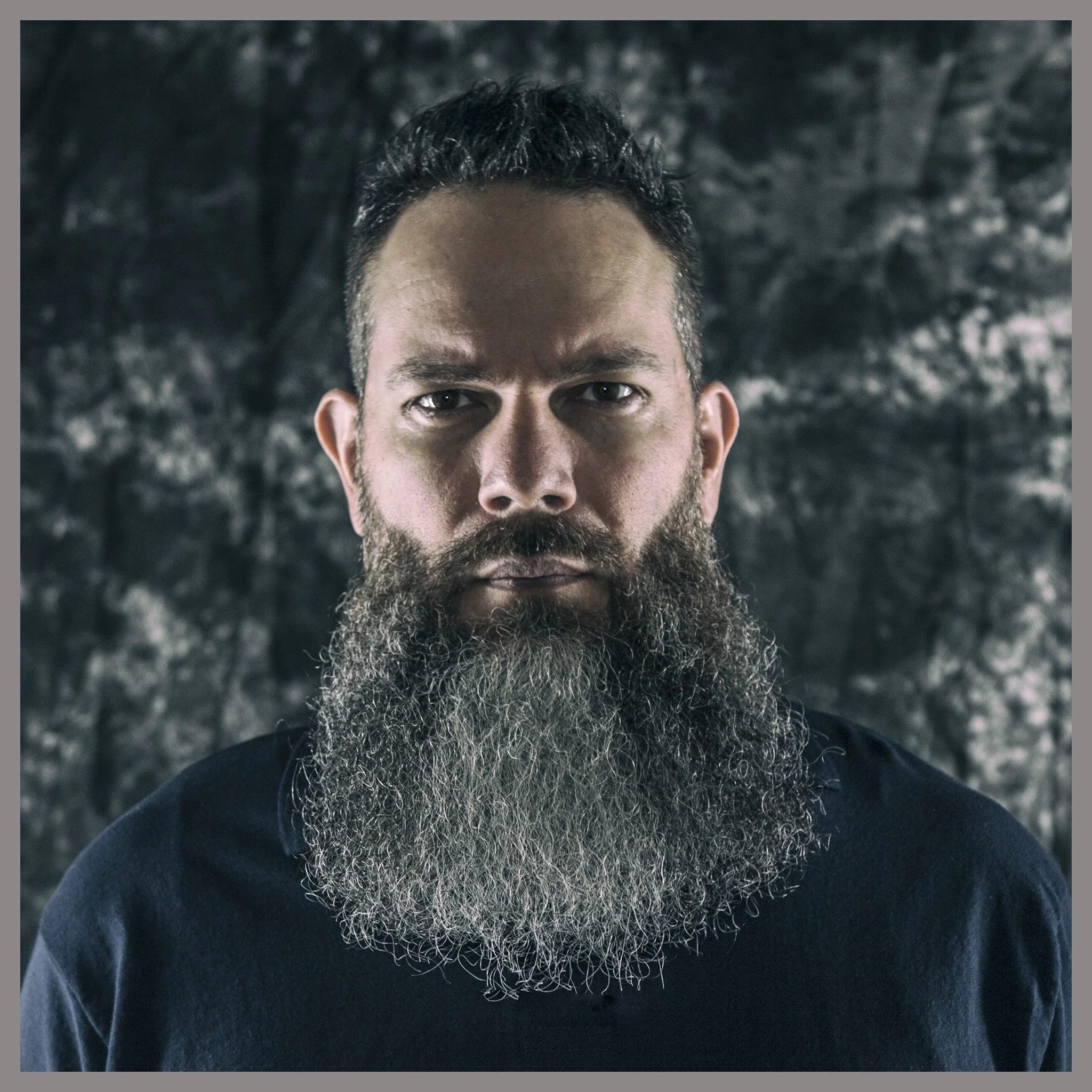 A man with a long, graying beard and short, dark hair looks directly at the camera against a dark, textured background.