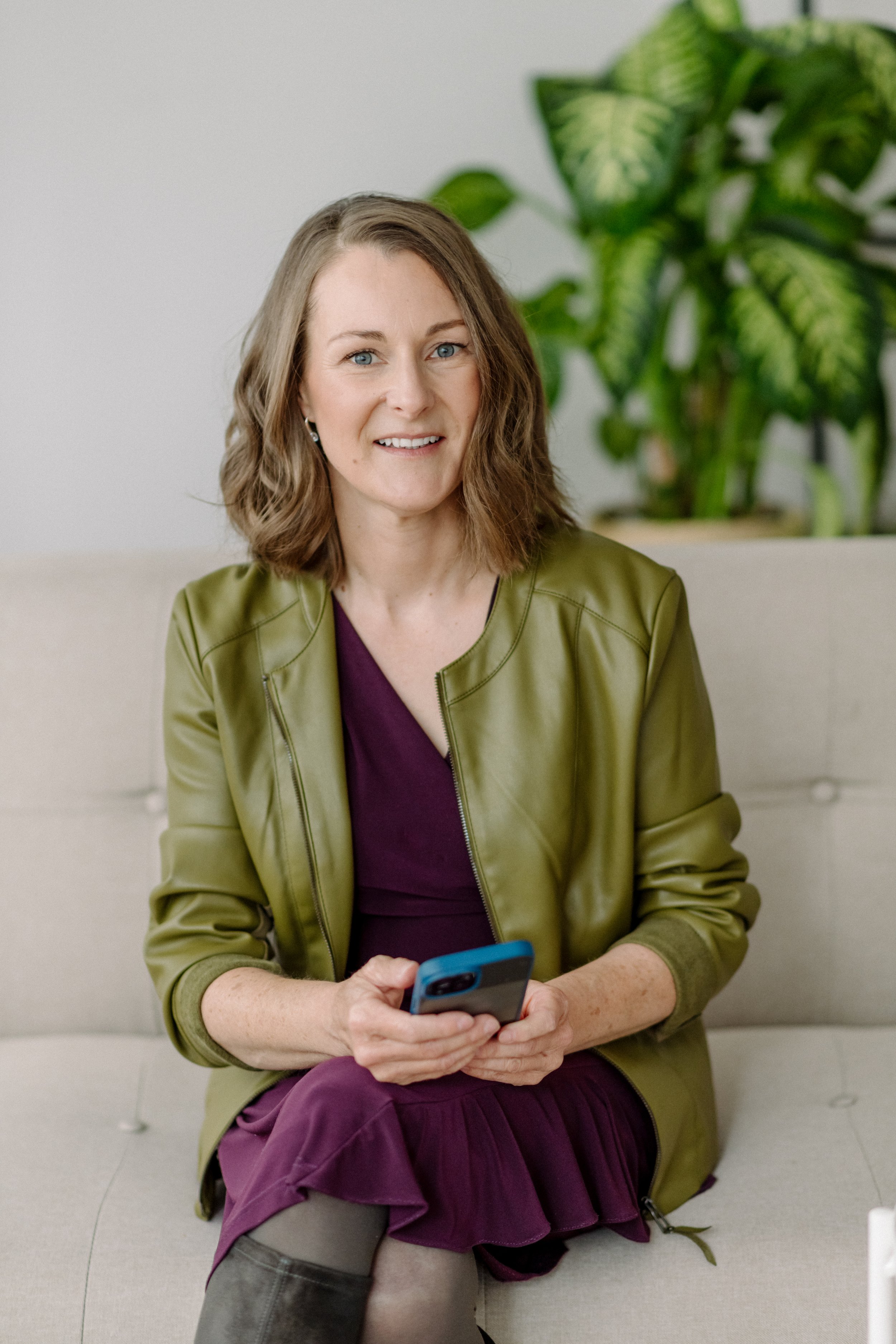 Eine lachende Frau mit blauen Augen, braunen Haaren und Ohrringen, die auf einem Sofa sitzt und ein Smartphone hält. Sie trägt eine grüne Jacke, eine dunkelviolette Bluse und einen schwarzen Rock. Im Hintergrund eine große grüne Pflanze.