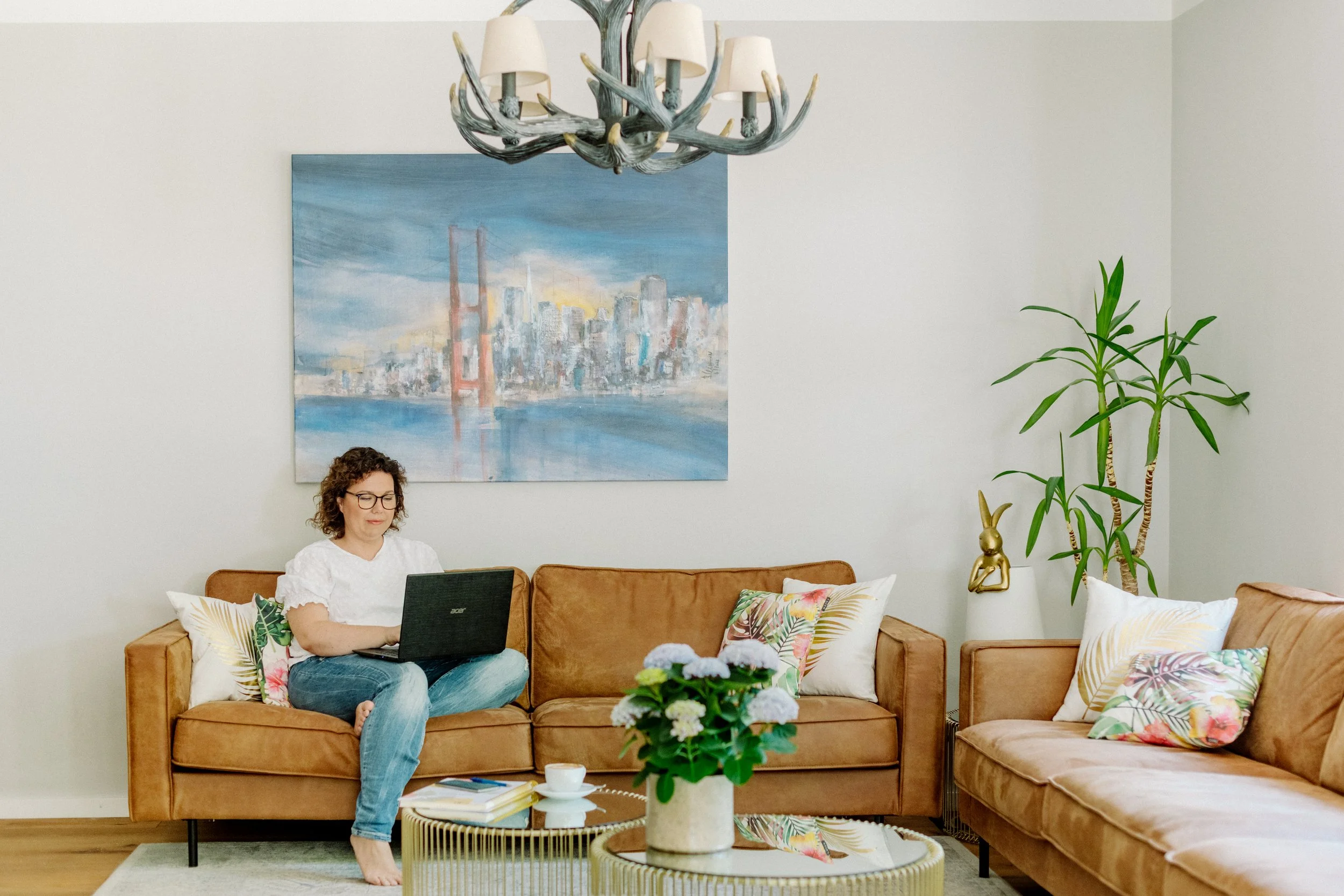 Eine Frau sitzt entspannt auf einem braunen Sofa mit bunten, pflanzen- und blumenmusternden Kissen, arbeitet an einem Laptop, im Wohnzimmer mit moderner Dekoration, darunter ein Gemälde einer Stadt und ein abstrakter Leuchter an der Decke, eine große