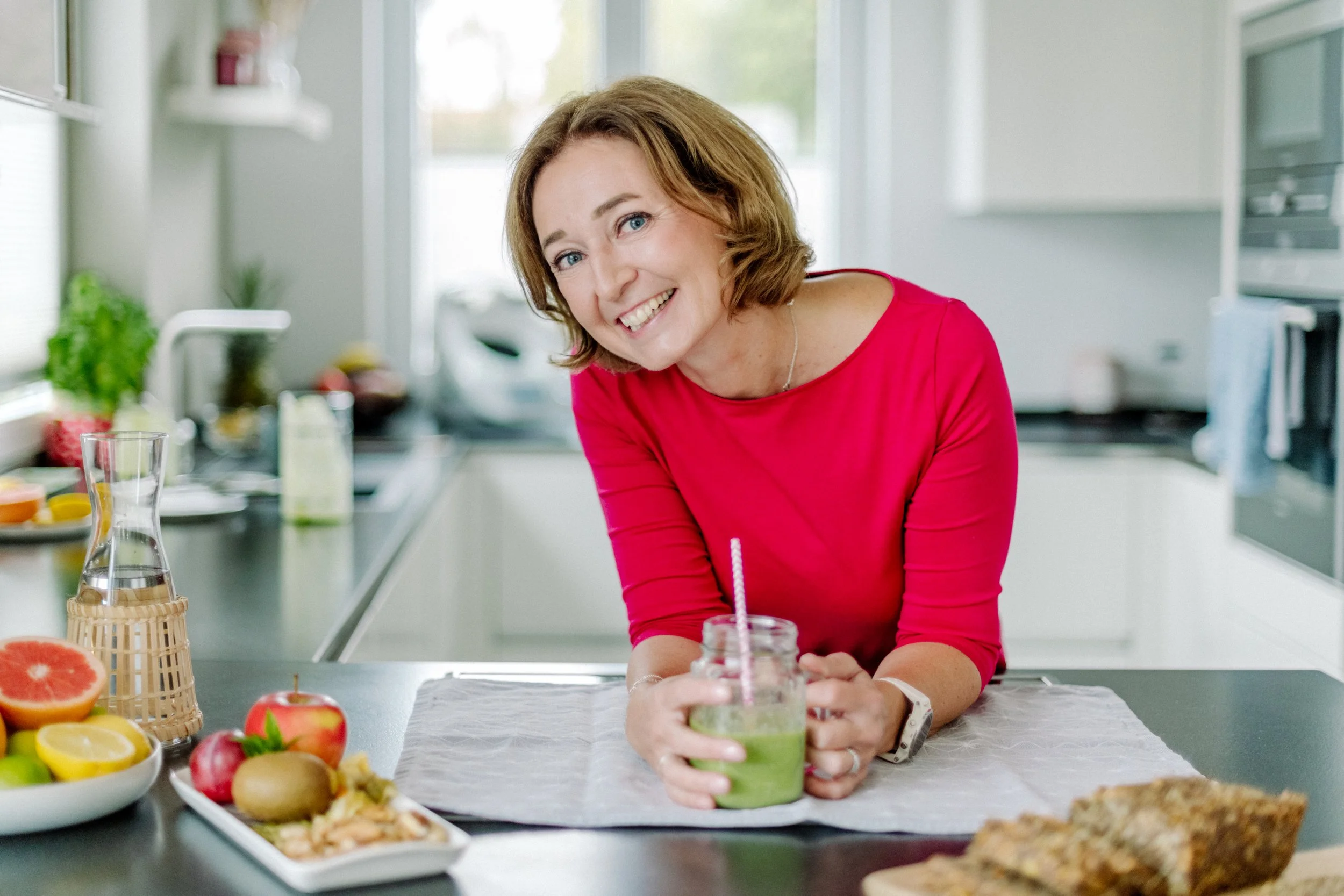 Eine lachende Frau in rotem Oberteil steht in einer modernen, hellen Küche und hält ein Glas mit grünen Smoothie mit Strohhalm. Der Tisch im Vordergrund ist mit frischem Obst und Speisen gedeckt.