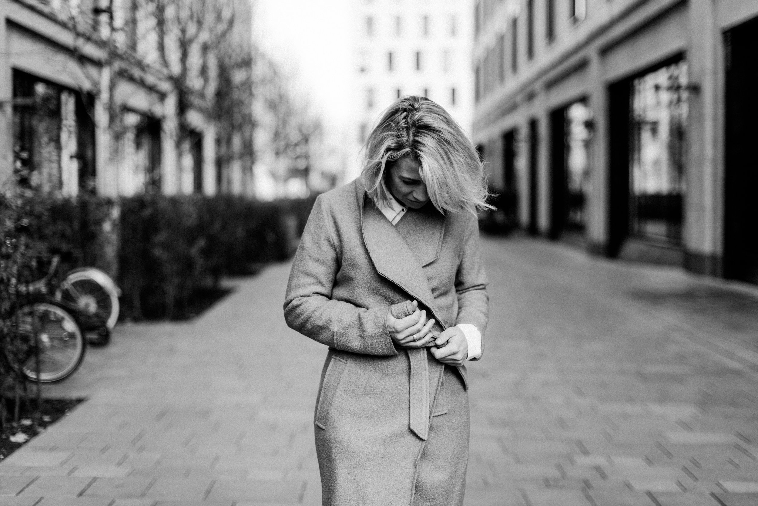 Eine Frau mit lockigem, blondem Haar trägt eine graue Jacke und einen grauen Rock, steht auf einem Gehweg in einer Stadt und schaut nach unten.