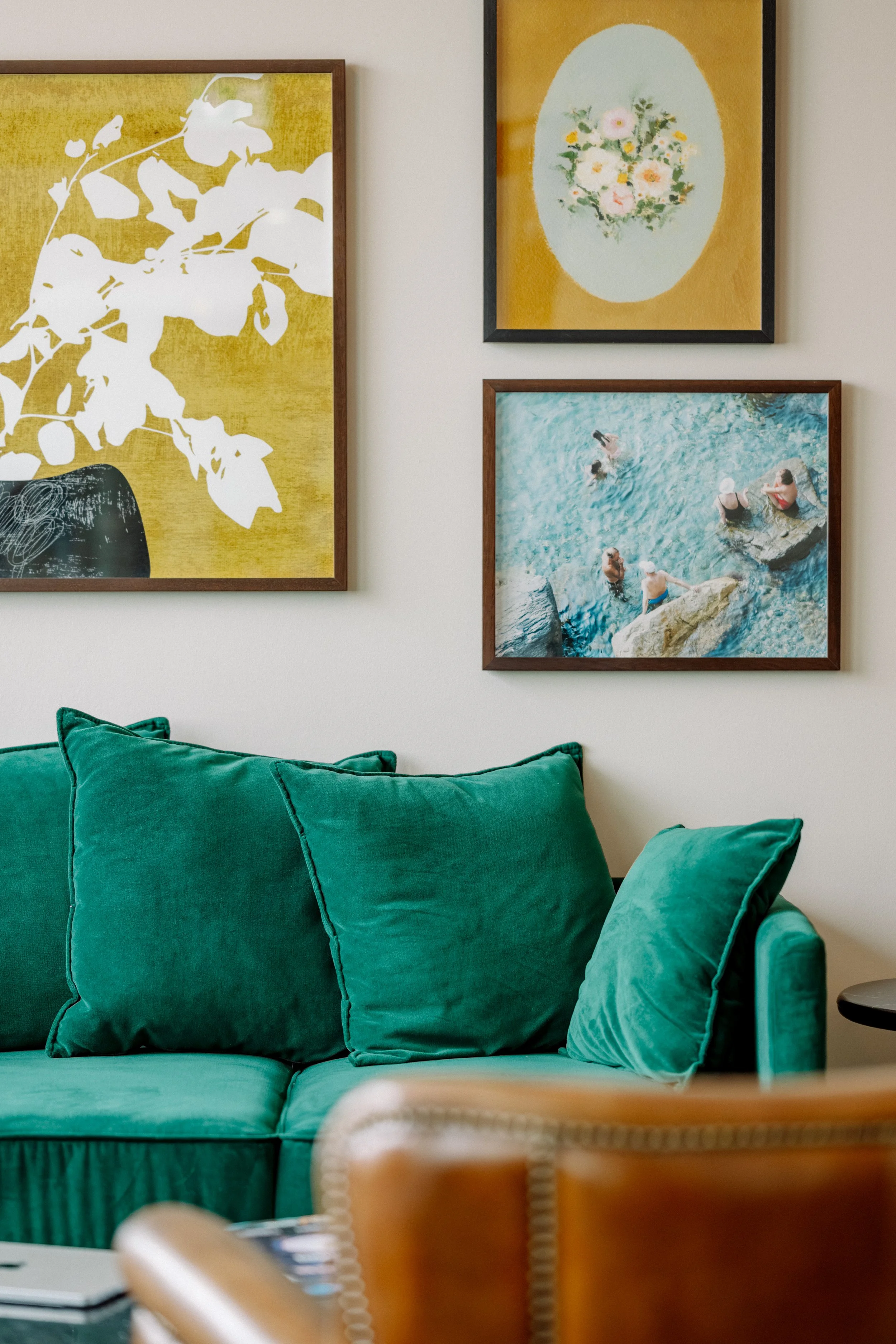 Ein Wohnzimmer mit grüner Couch und mehreren grünen Kissen, Wandbilder mit Blumen und Wasser, ein Holzsessel im Vordergrund.