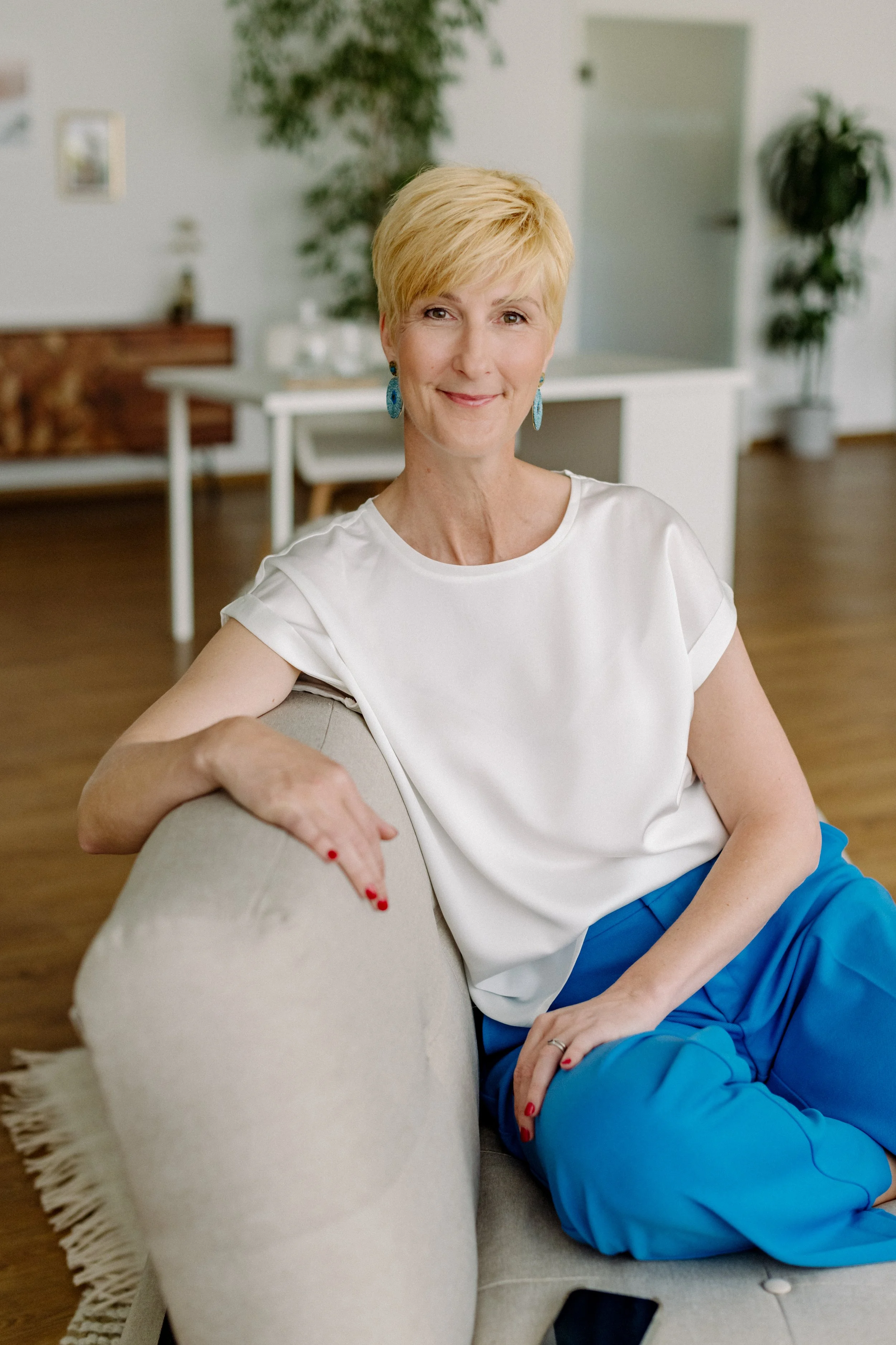 Eine lächelnde ältere Frau mit kurzen blonden Haaren sitzt entspannt auf einem Sofa in einem modern eingerichteten Wohnzimmer, trägt eine weiße Bluse, blaue Hosen und auffällige blaue Ohrringe.