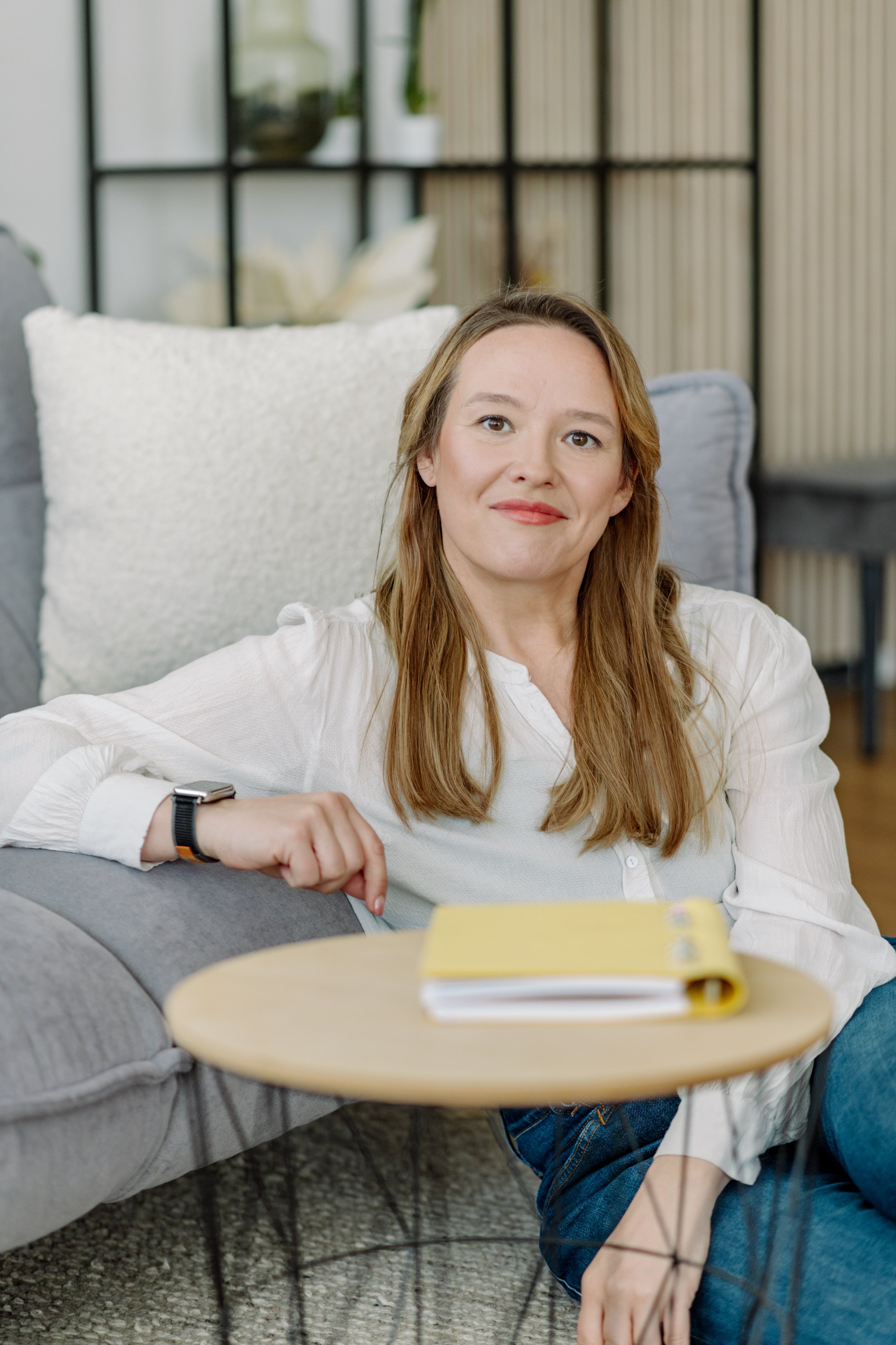 Junge Frau sitzt auf einem Sofa, liest ein Buch und lächelt, im Hintergrund ein Bücherregal.