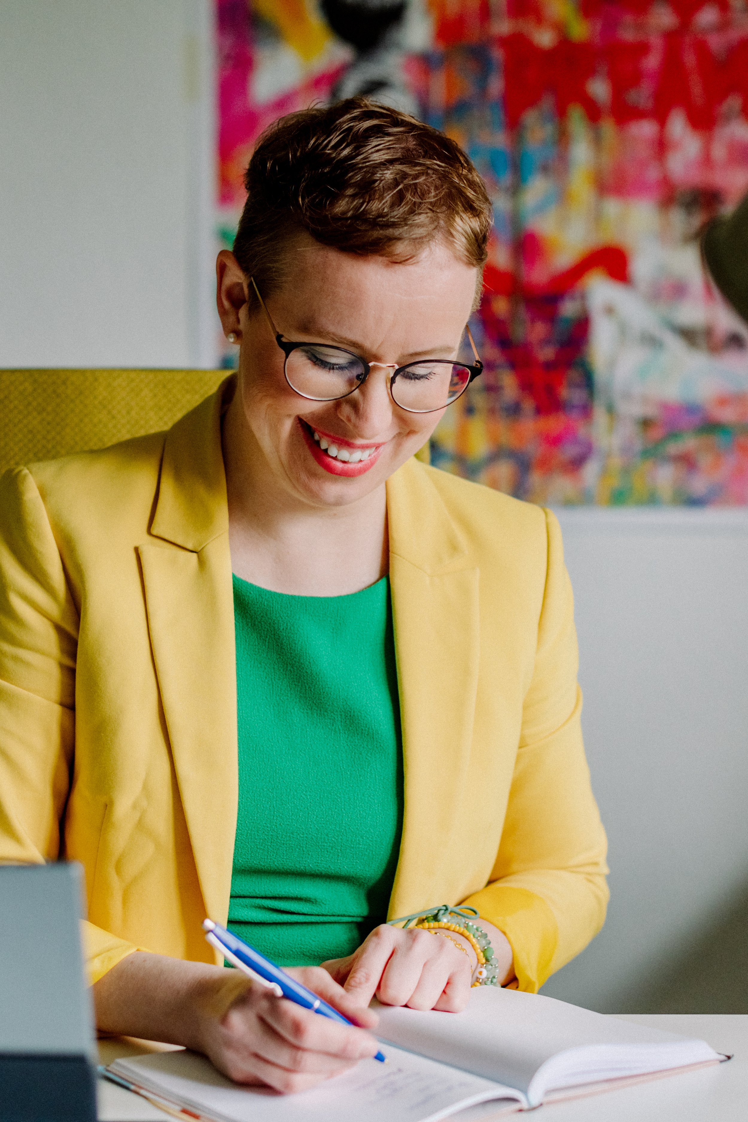 Eine Frau mit kurzen braunen Haaren, die eine gelbe Jacke und ein grünes Oberteil trägt, arbeitet an einem Notizbuch und hält einen blauen Kugelschreiber. Im Hintergrund befindet sich eine bunte abstrakte Wandkunst.