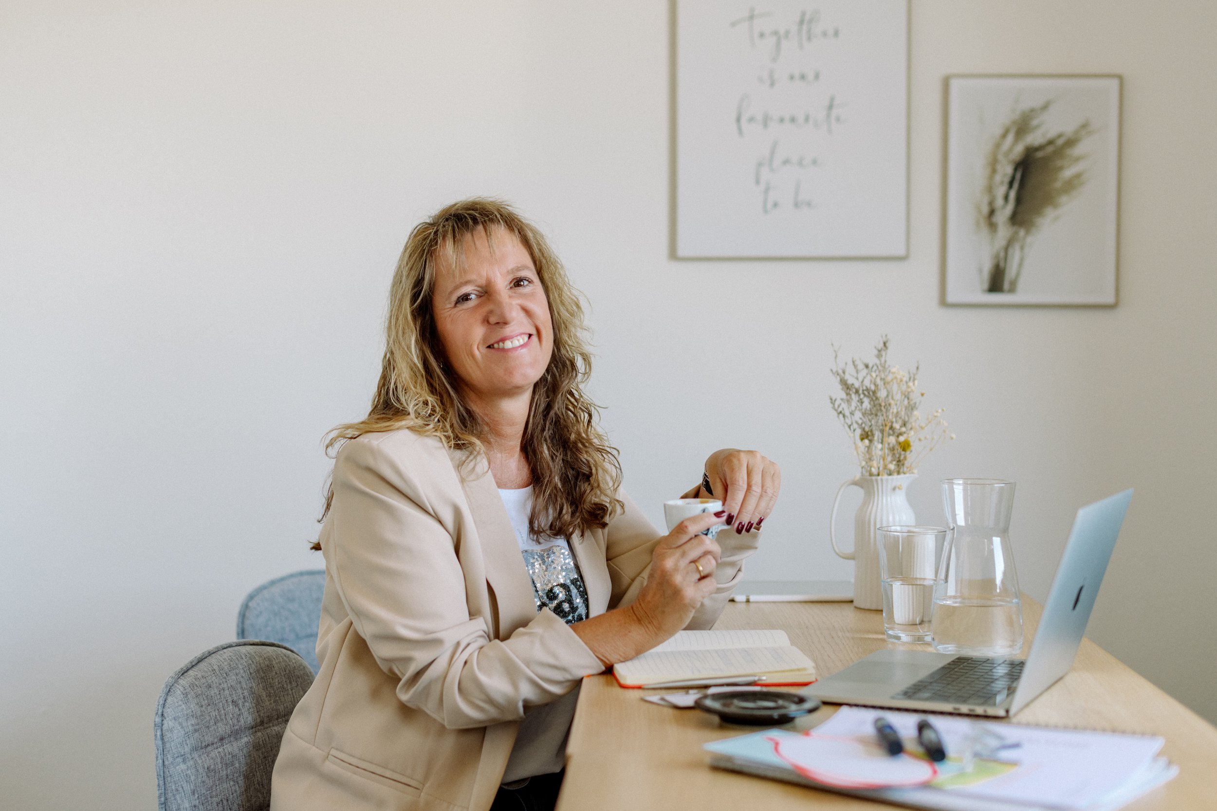 Eine Frau mit lockigem, schulterlangem Haar sitzt an einem Tisch, trinkt aus einer Tasse und hat einen Laptop vor sich. Im Hintergrund sind weiße Wände mit Kunst und einer Notiz. Auf dem Tisch stehen Gläser mit Wasser, Notizen, ein Notizbuch, Stifte und eine Pflanze.
