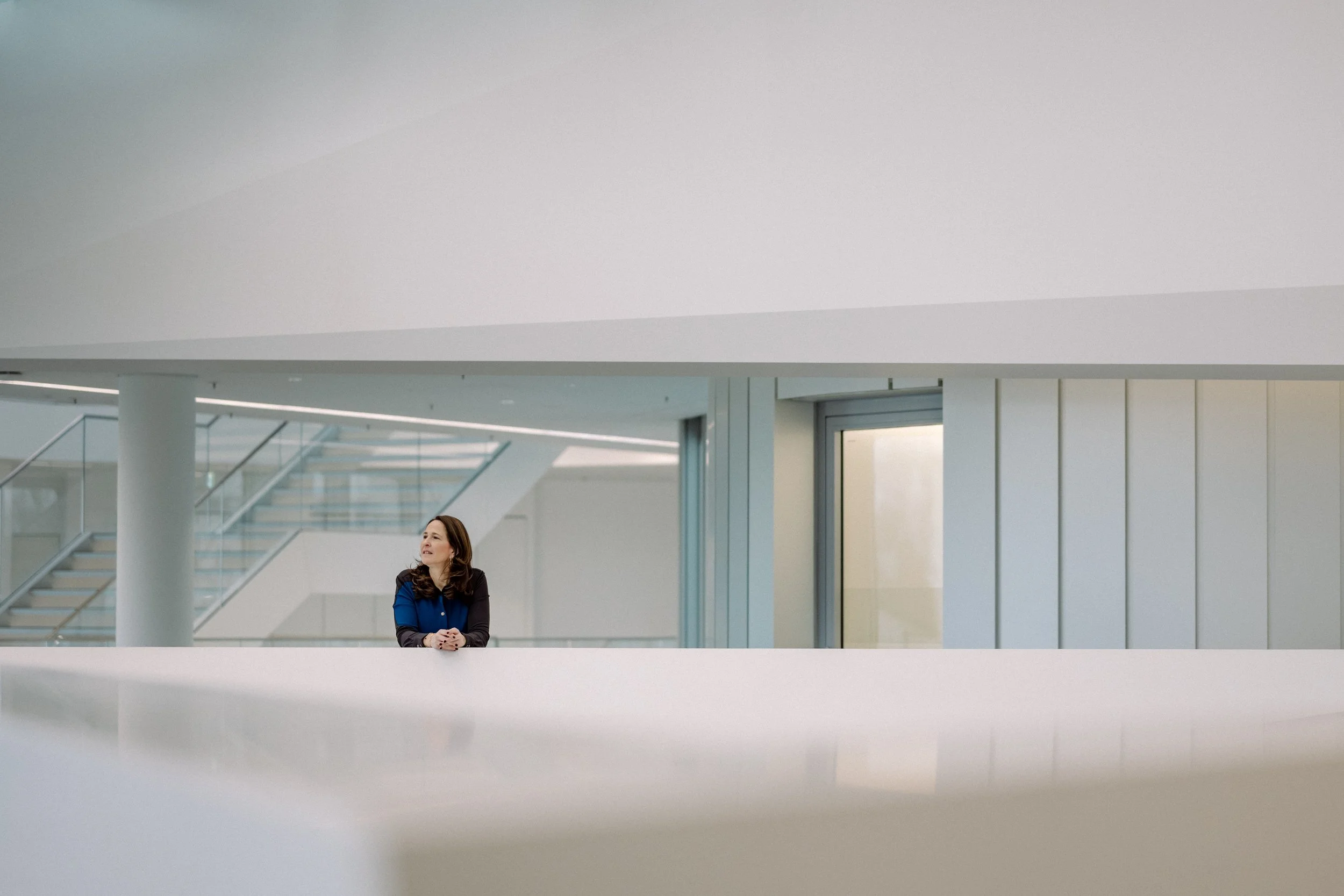 Eine Frau steht in einem modernen, hellen Gebäude mit großen Fenstern und Treppen, sie trägt einen blauen und schwarzen Blazer.