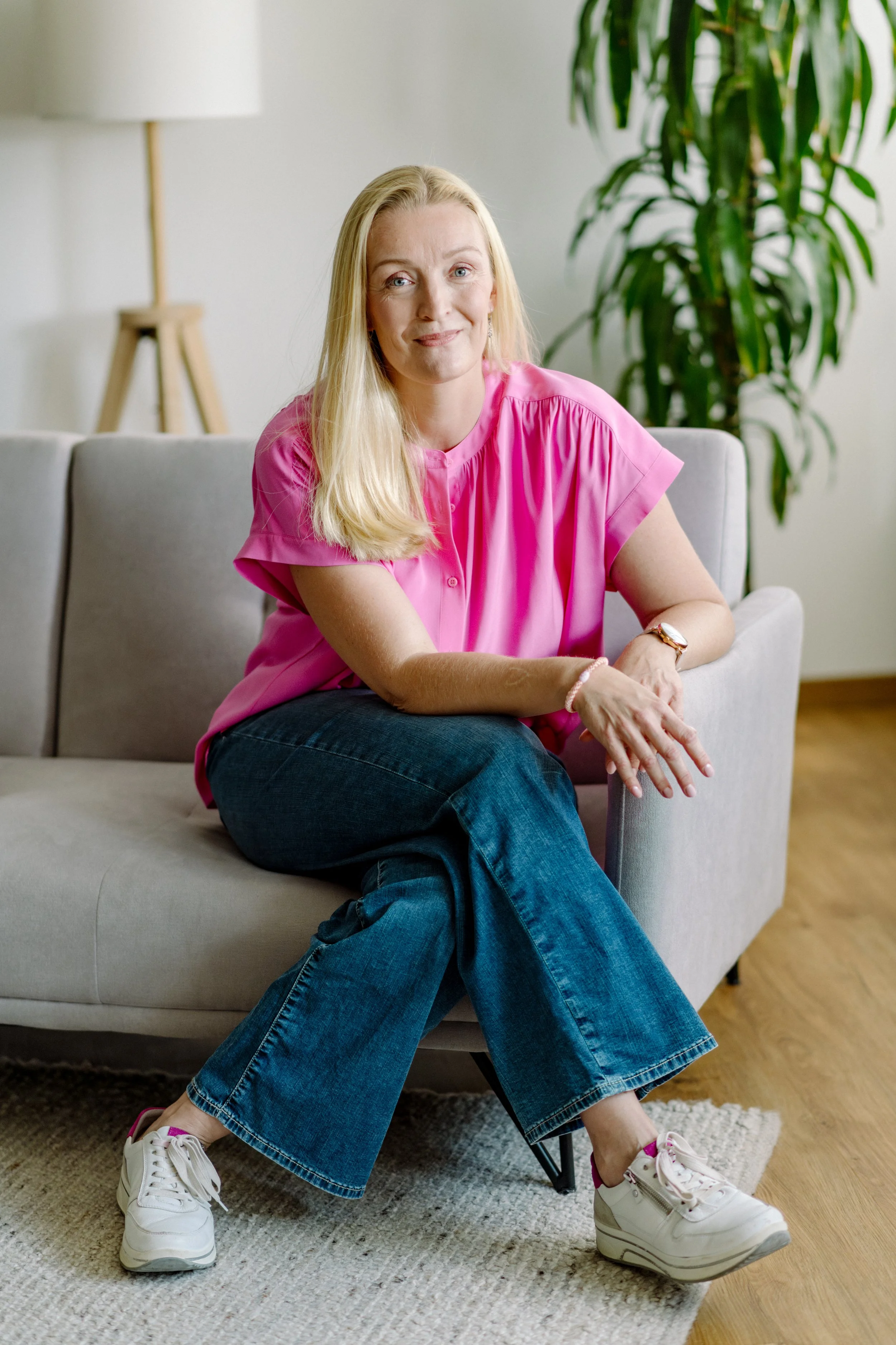 Eine blondierte Frau in einer rosa Bluse sitzt auf einem grauen Sofa im Wohnzimmer, neben ihr eine große Zimmerpflanze, sie lächelt sanft.