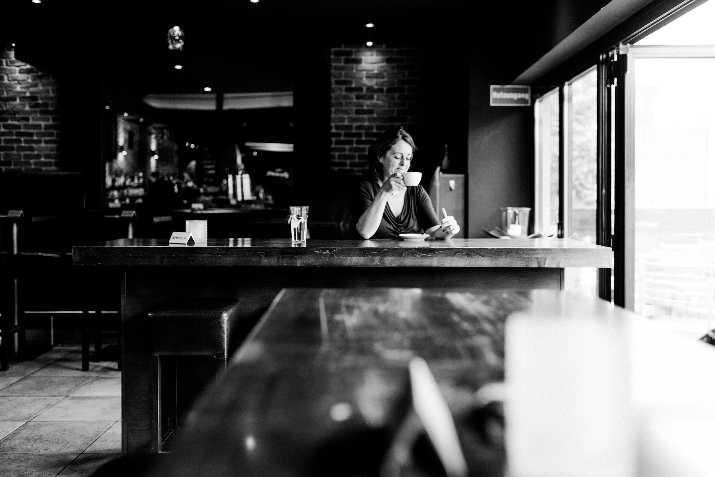 Eine Frau sitzt in einem Café, trinkt aus einer Tasse und schaut aus dem Fenster, im Hintergrund ist eine Bar und ein Fenster, das Tageslicht hereinlässt. Das Bild ist schwarz/weiß.