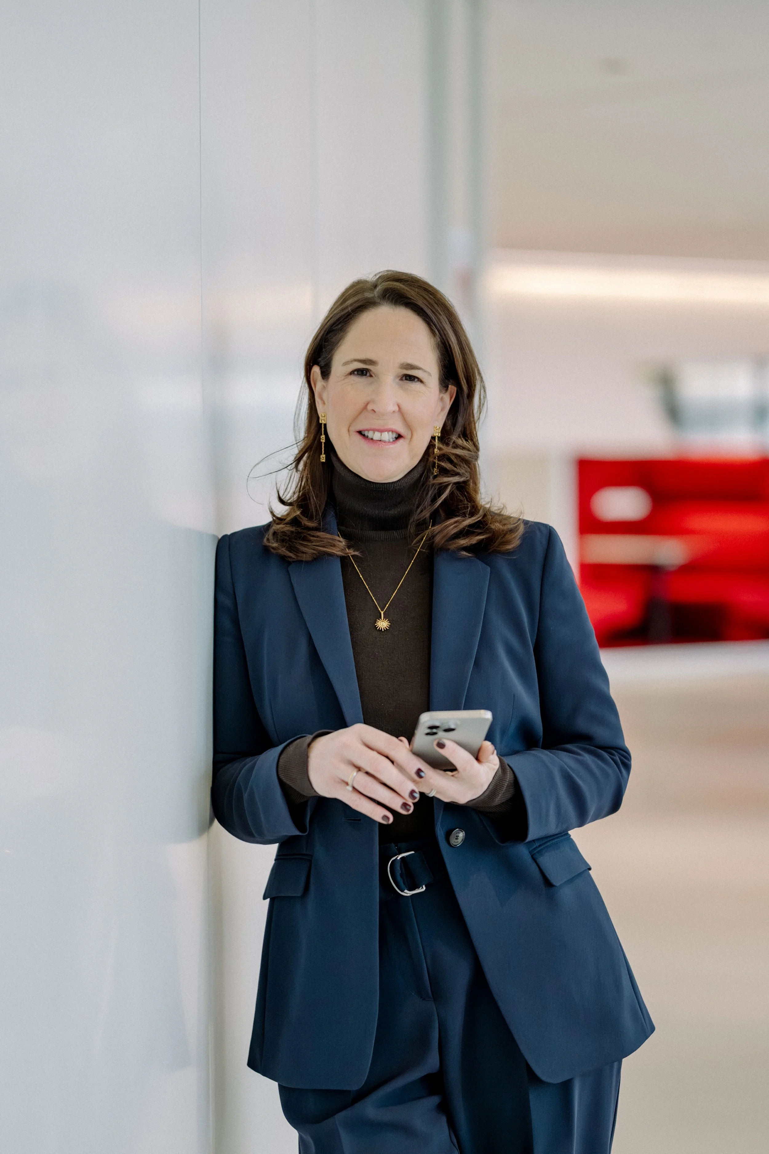 Eine Frau im Business-Outfit steht in einem modernen Büro, lehnt an einer weißen Wand und hält ein Smartphone in der Hand, lächelt freundlich.