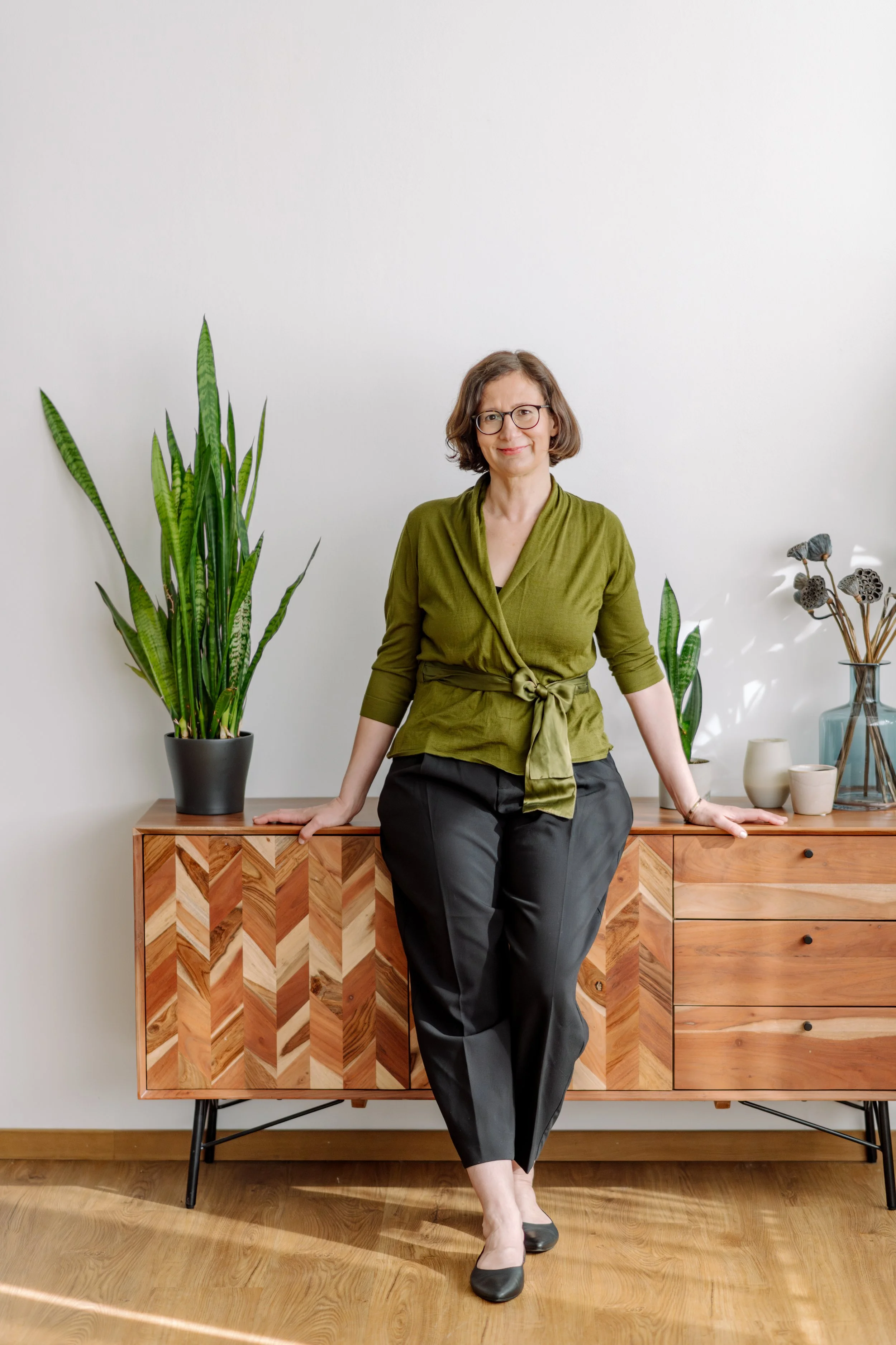 Eine Frau mit Brille sitzt auf einer Holzkommode vor weißer Wand, umgeben von Grünpflanzen und Keramikvasen.