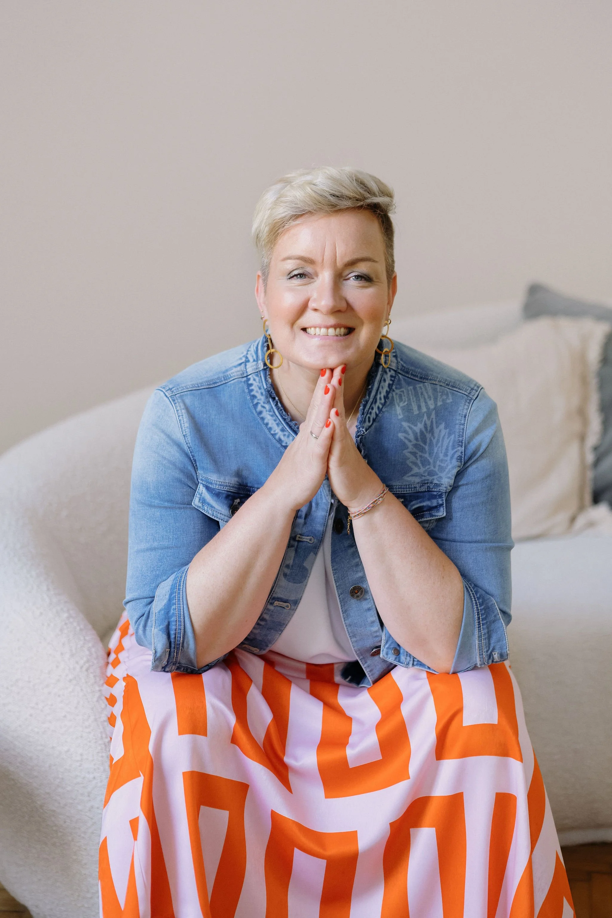 Eine lachende Frau mit kurzen grauen Haaren, die in einem Strand- oder Sommer-Outfit sitzt, trägt eine goldene Creole-Ohrringe, eine Jeansjacke und ein orange-weiß gestreiftes Kleid. Sie sitzt auf einem Sofa oder Stuhl vor einem neutralen Hintergrund.