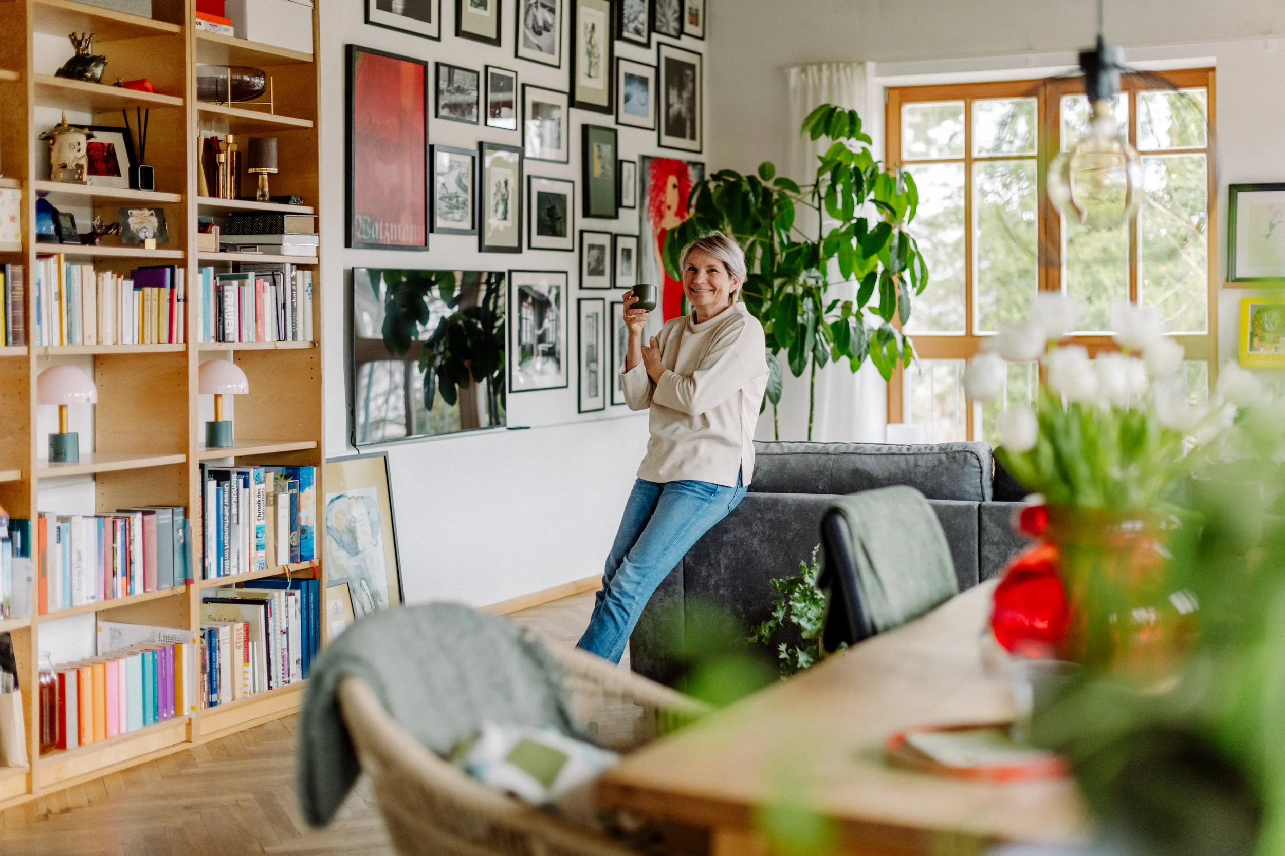 Eine ältere Frau steht in einem Wohnzimmer, hält eine Tasse und lächelt. Das Wohnzimmer ist mit vielen Bildern an der Wand, einem großen grünen Pflanze, einem Fernseher und einem Bücherregal dekoriert. Im Vordergrund sind unscharf Blumen auf einem Ti