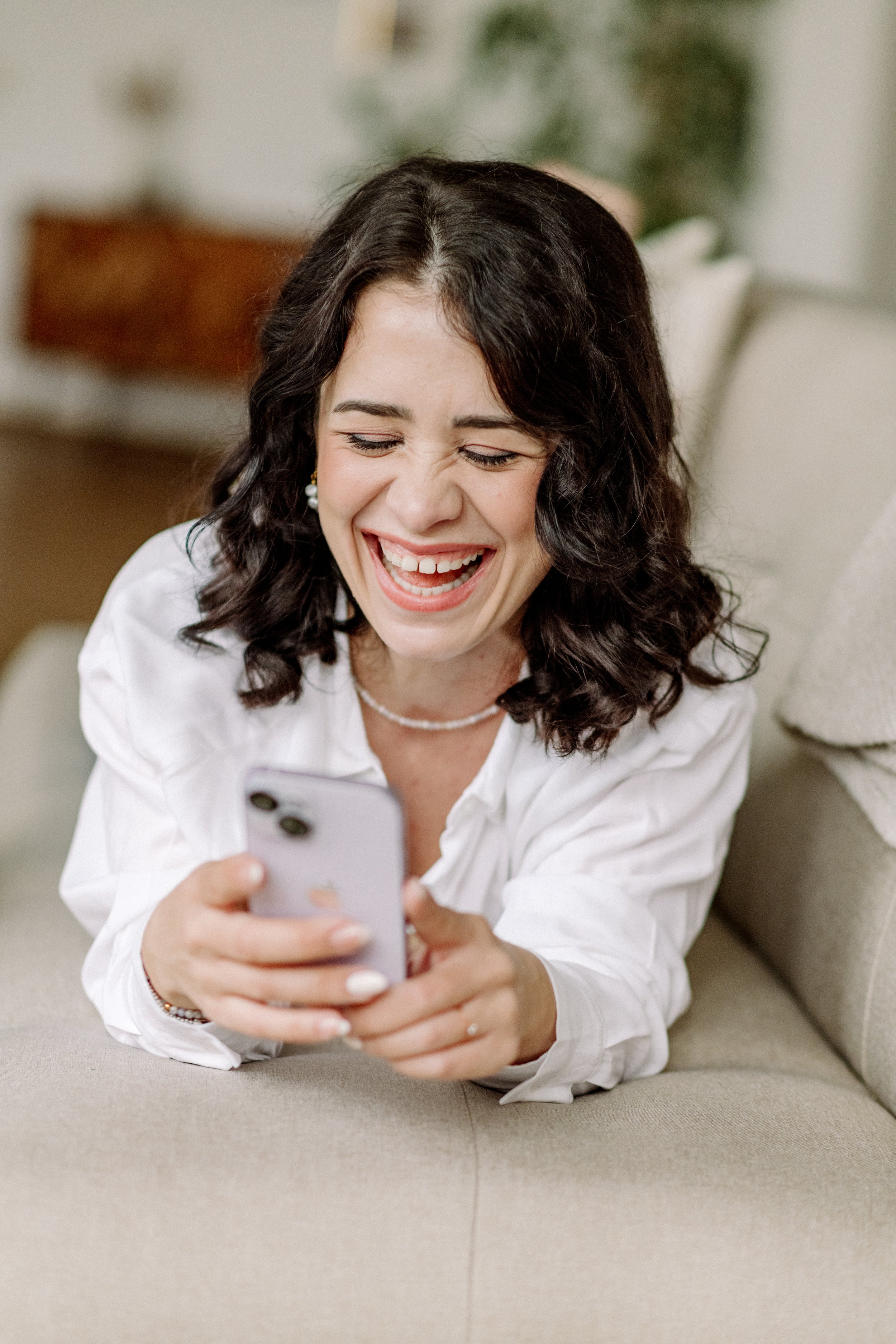 Junge Frau mit braunen, gelockten Haaren, lächelnd und lachend, zeigt auf ihr Smartphone, während sie auf einem Sofa sitzt.