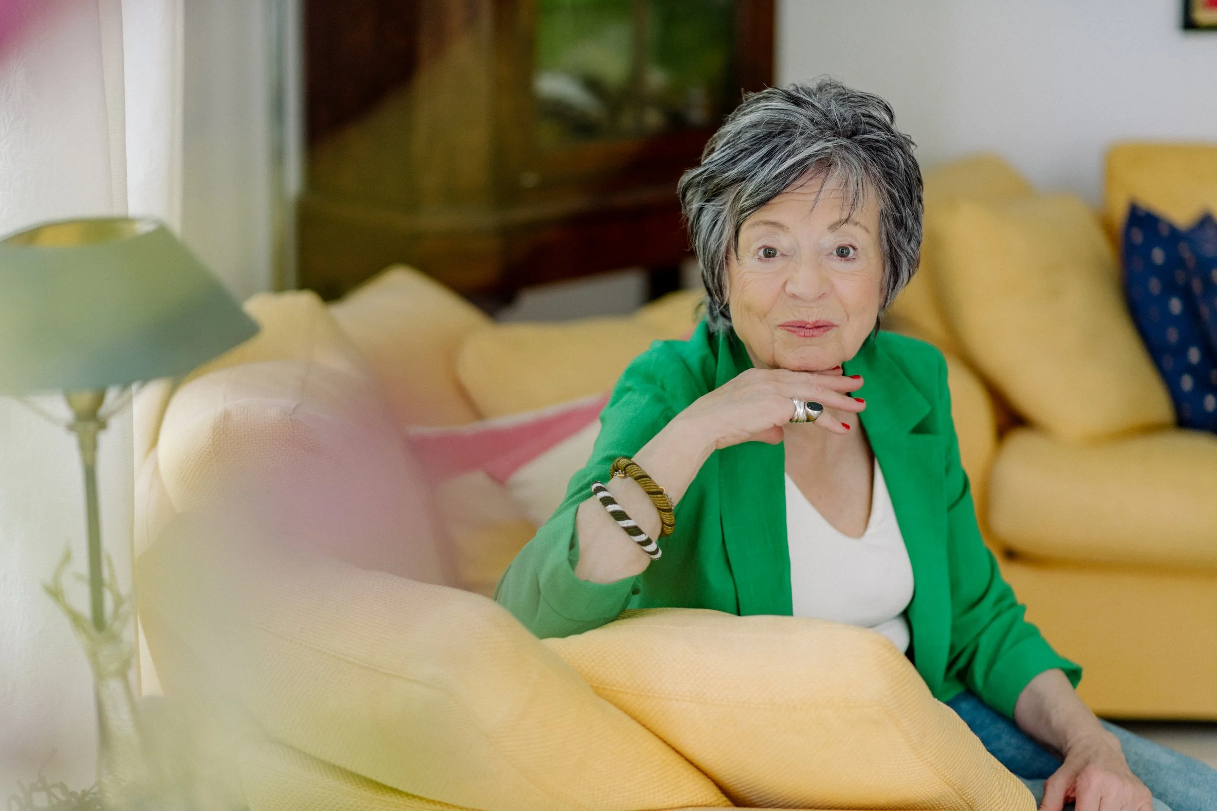 Eine ältere Frau mit grau-schwarz gesprenkeltem Haar sitzt auf einem gelben Sofa, trägt eine grüne Jacke und schaut nach oben, während sie ihre Hand an ihr Kinn legt.
