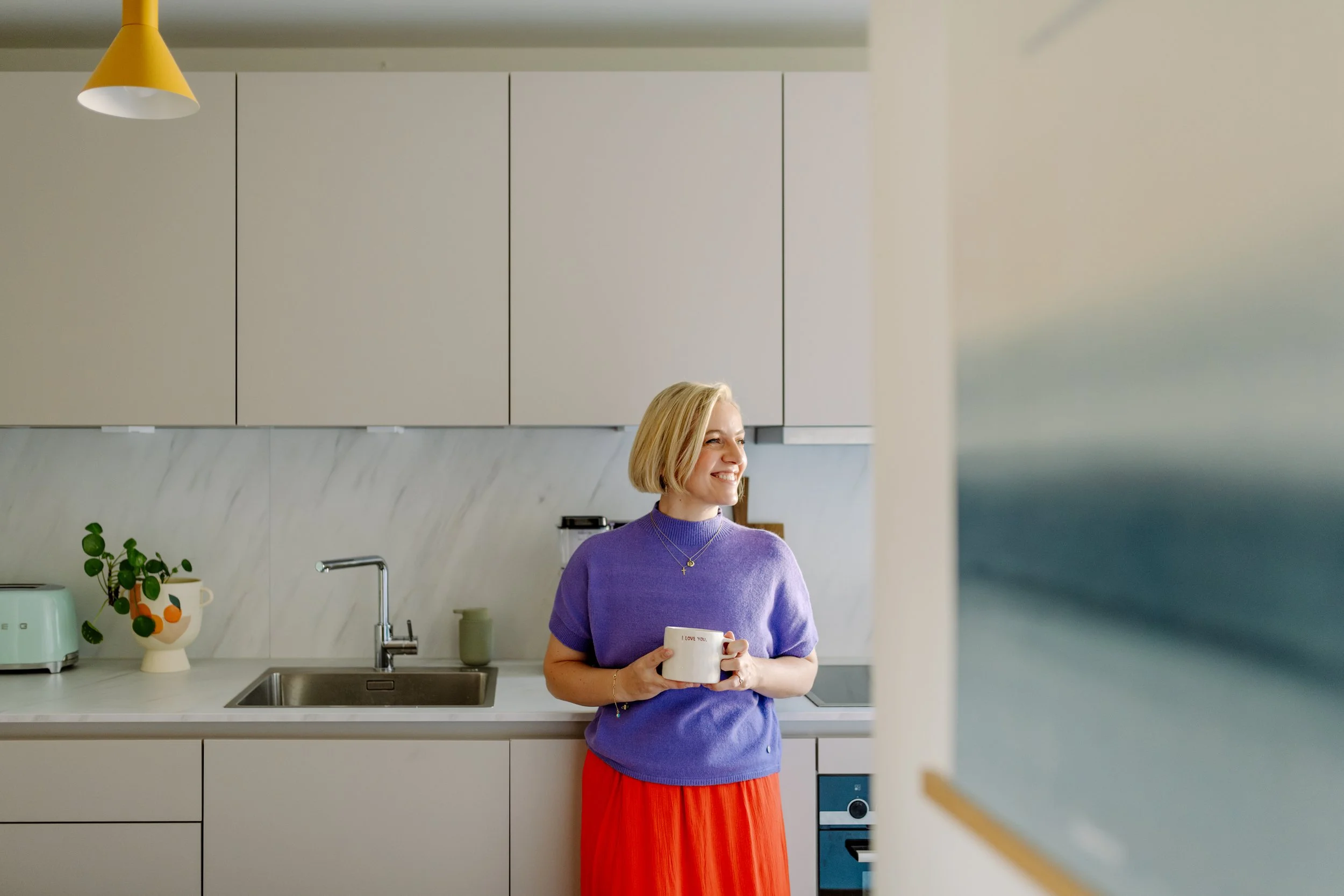 Eine lachende Frau mit blonden Haaren in einem lilafarbenen Pullover hält eine weiße Tasse mit der Aufschrift 'I love you' in einer modernen Küche.