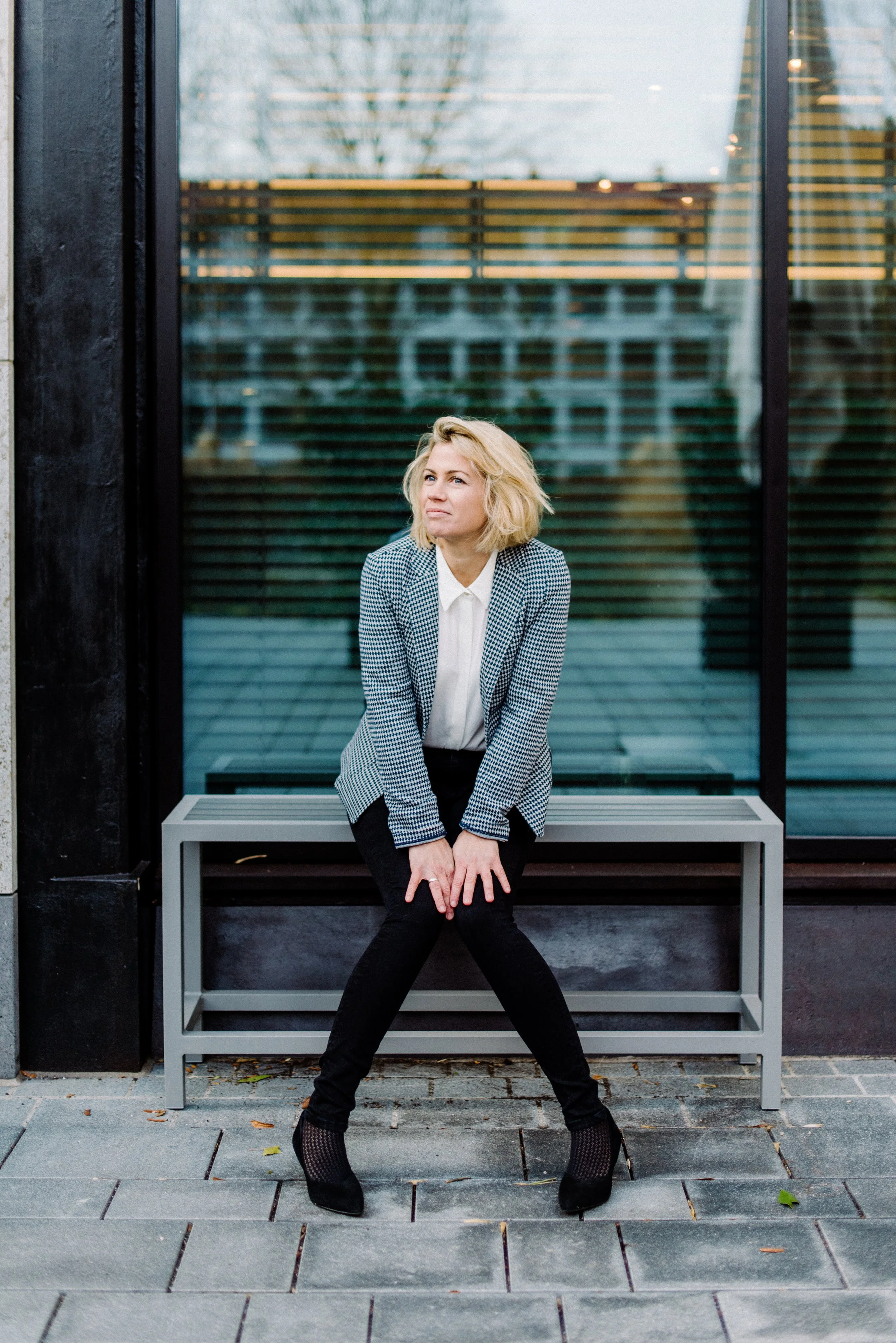 Eine Frau mit blonden, lockigen Haaren in einem grauen und weißen karierten Blazer sitzt auf einer Bank vor einem modernem Gebäude mit großen Fenstern und horizontalen Lamellen. Sie trägt schwarze Schuhe und schwarze Hose, und blickt nach oben.