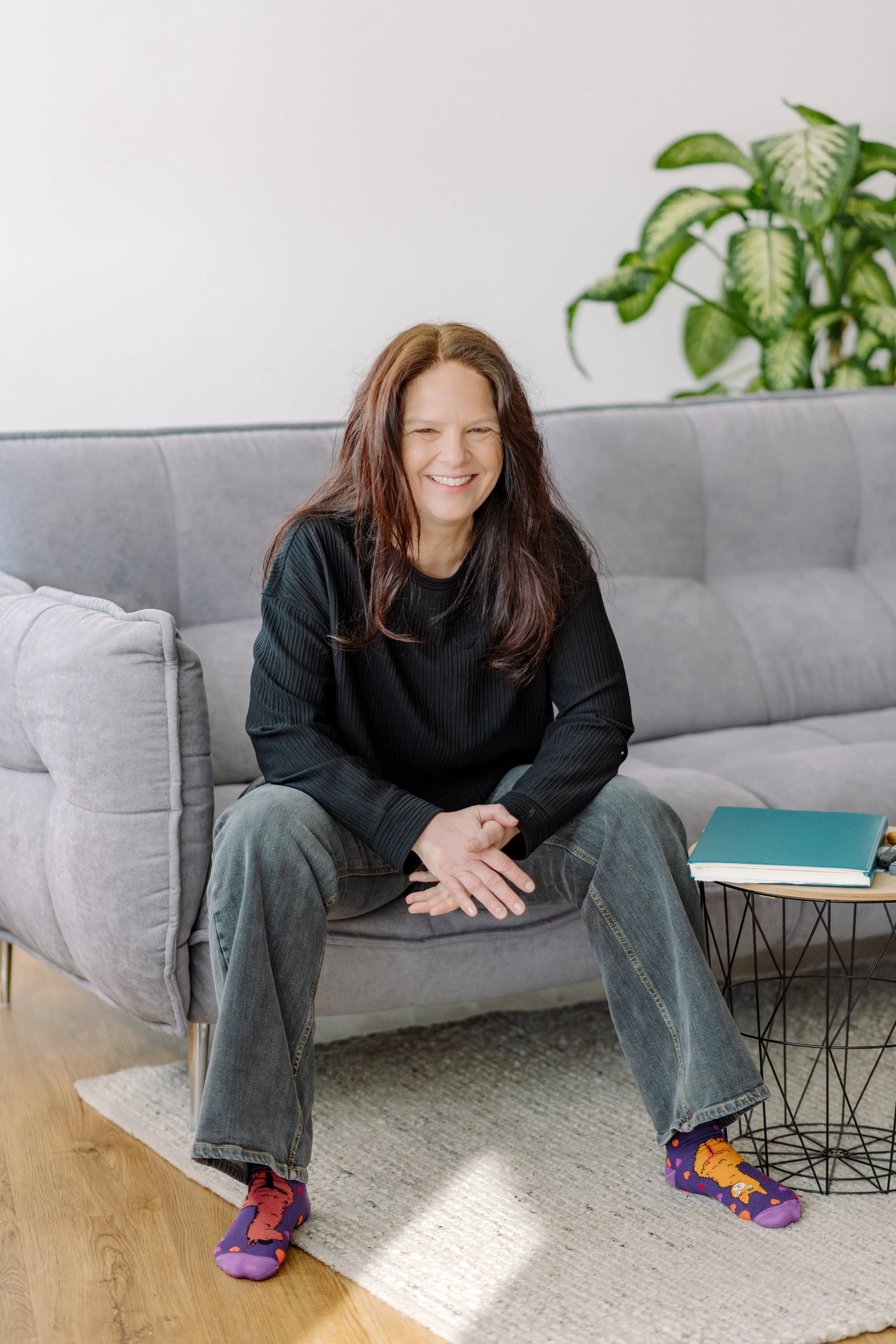 Eine lächelnde Frau sitzt auf einem grauen Sofa, trägt bunte Socken mit Tieren und Jeans, neben einem kleinen Beistelltisch mit einem blauen Buch.