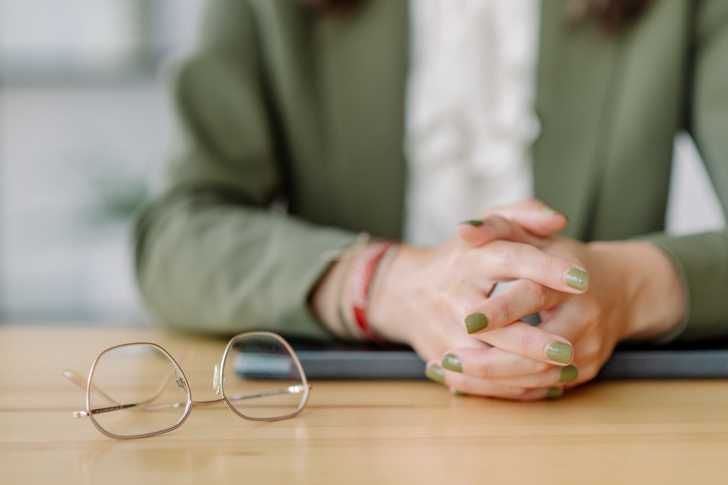 Nahaufnahme von fertigen Gold-Ohrringen auf einem Tisch, im Hintergrund eine Person mit gefalteten Händen, die grünen Nagellack trägt, und eine Brille liegt ebenfalls auf dem Tisch.