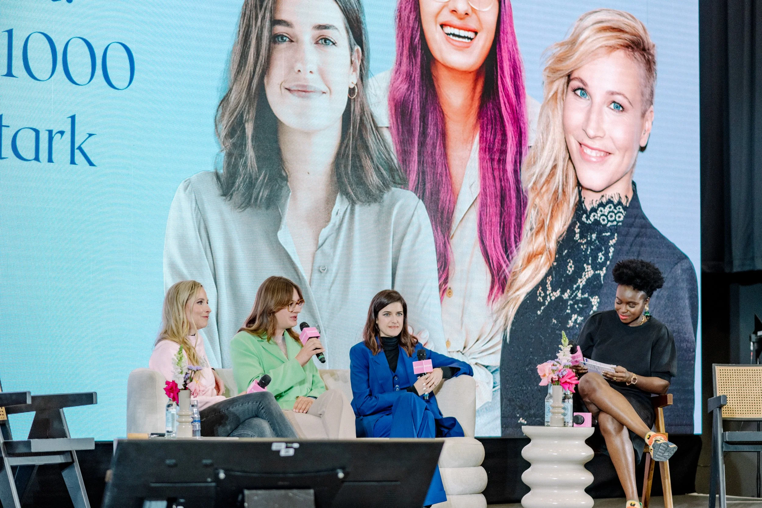 Eine Panel-Diskussion mit mehreren Frauen, die auf einer Bühne sitzen, einige sprechen in Mikrofone, mit Blumen und Wasserflaschen vor ihnen, während im Hintergrund ein großes Bildschirm mit Gesichtsbildern von Frauen zu sehen ist.