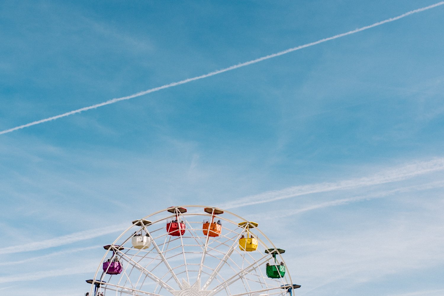 Ein Riesenrad mit bunten Gondeln vor einem blauen Himmel mit Wolken und -streifen.