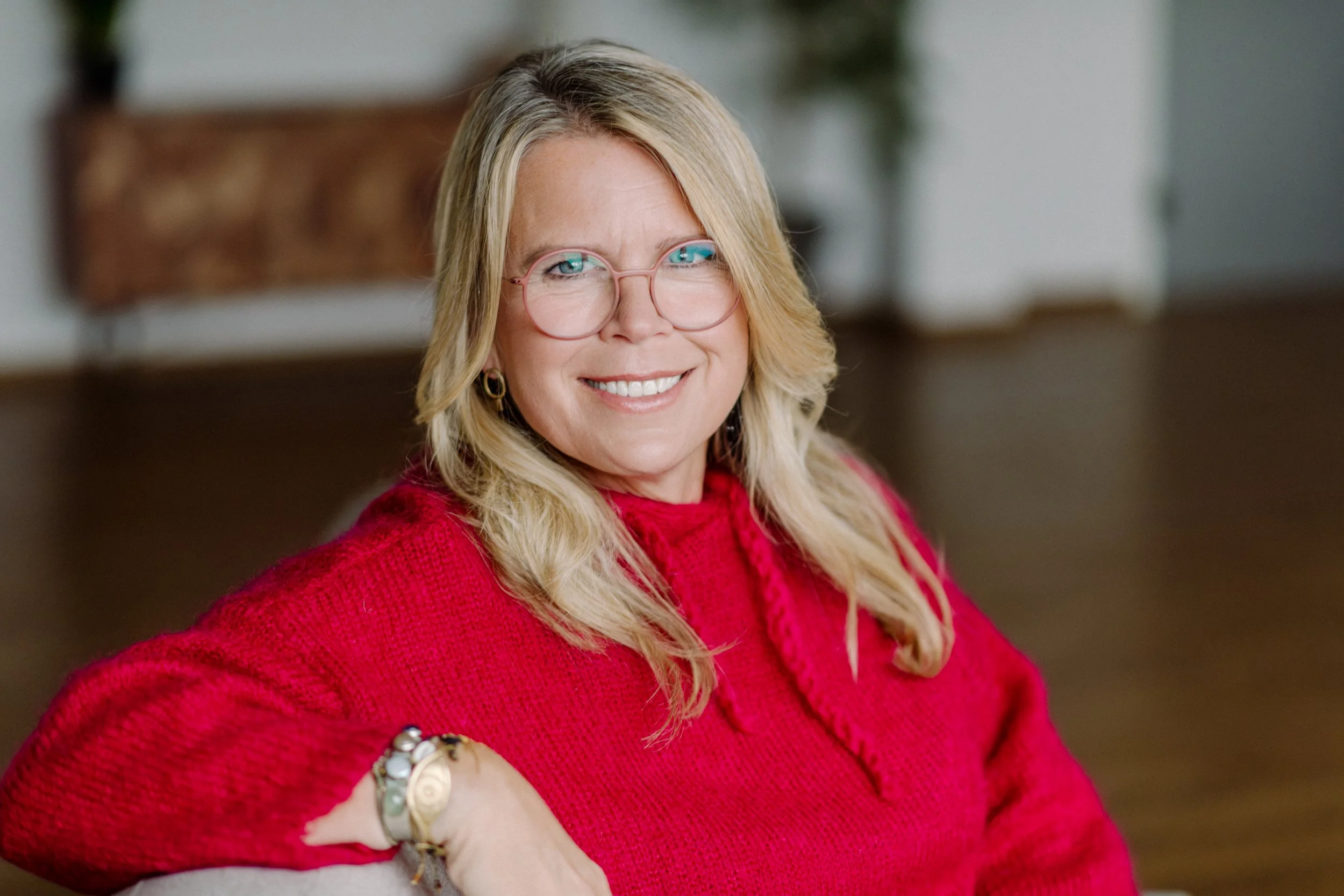 Eine lächelnde Frau mit langen blonden Haaren, trägt eine rosa Brille und eine rote Pullover, sitzt in einem gemütlichen Raum.
