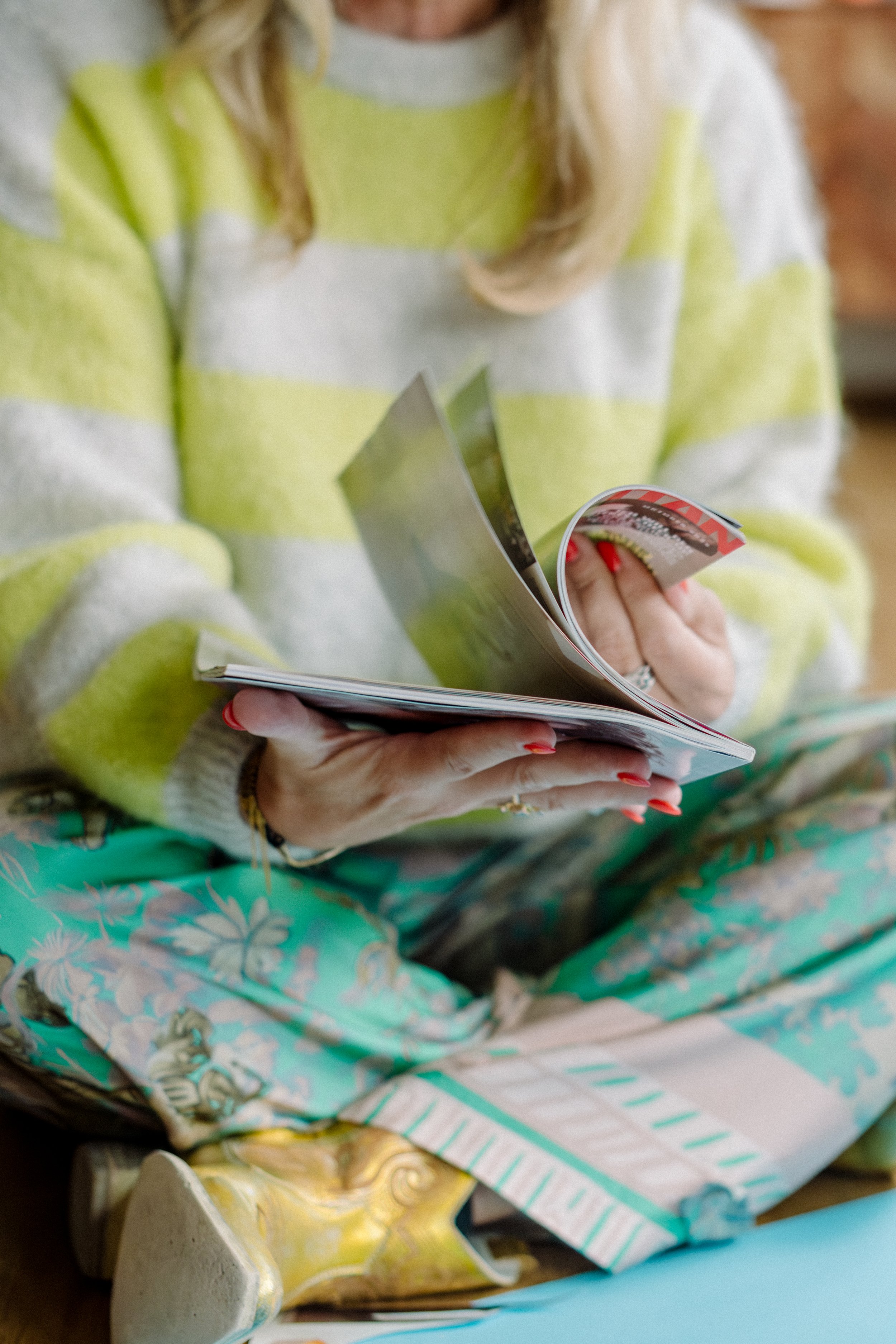 Eine Person hält ein aufgeschlagenes Magazin in den Händen, während sie auf einer bunten, floralen Decke sitzt.