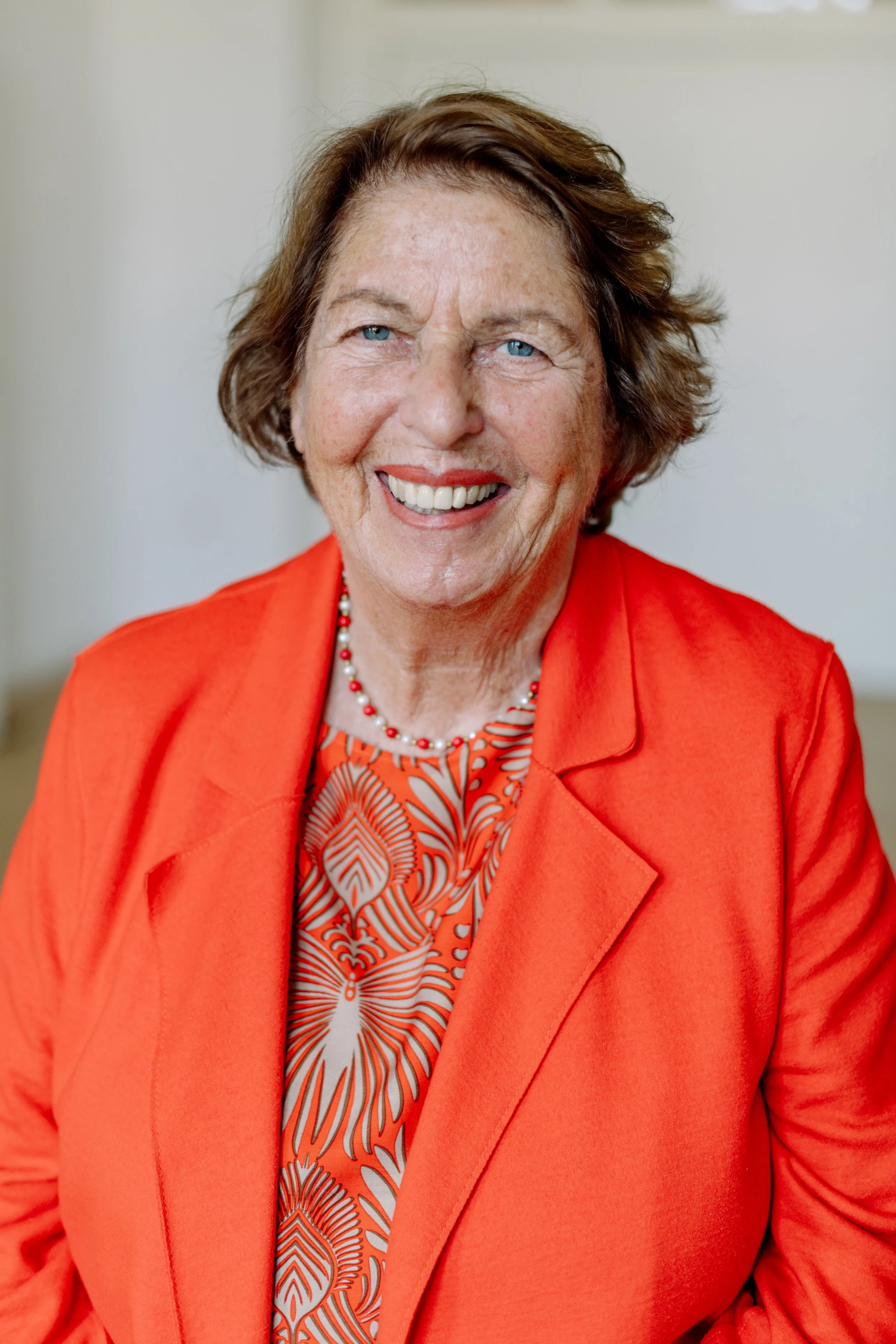 Eine älterer Frau mit kurzen braunen Haaren, blauen Augen und einem freundlichen Lächeln. Sie trägt eine orange Jacke und eine Perlenkette. Der Hintergrund ist neutral.
