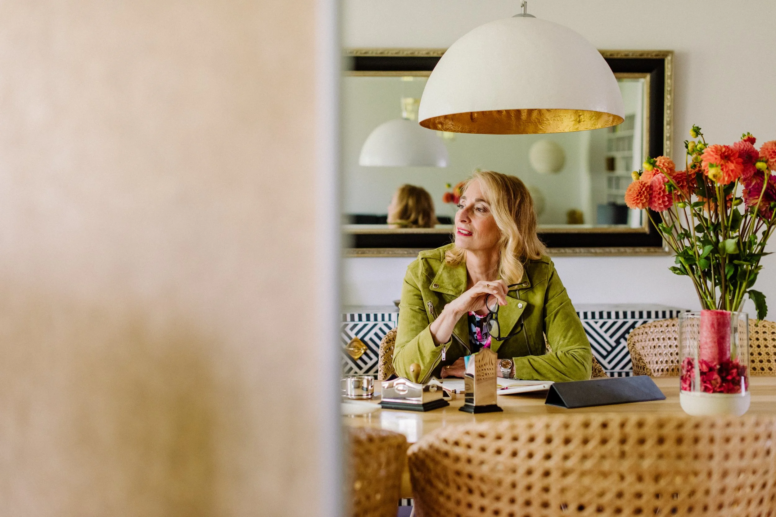 Eine blonde Frau sitzt an einem Tisch in einem Restaurant oder Café, umgeben von Blumenvasen, mit einem Spiegel im Hintergrund. Sie trägt eine grüne Jacke und lächelt leicht.