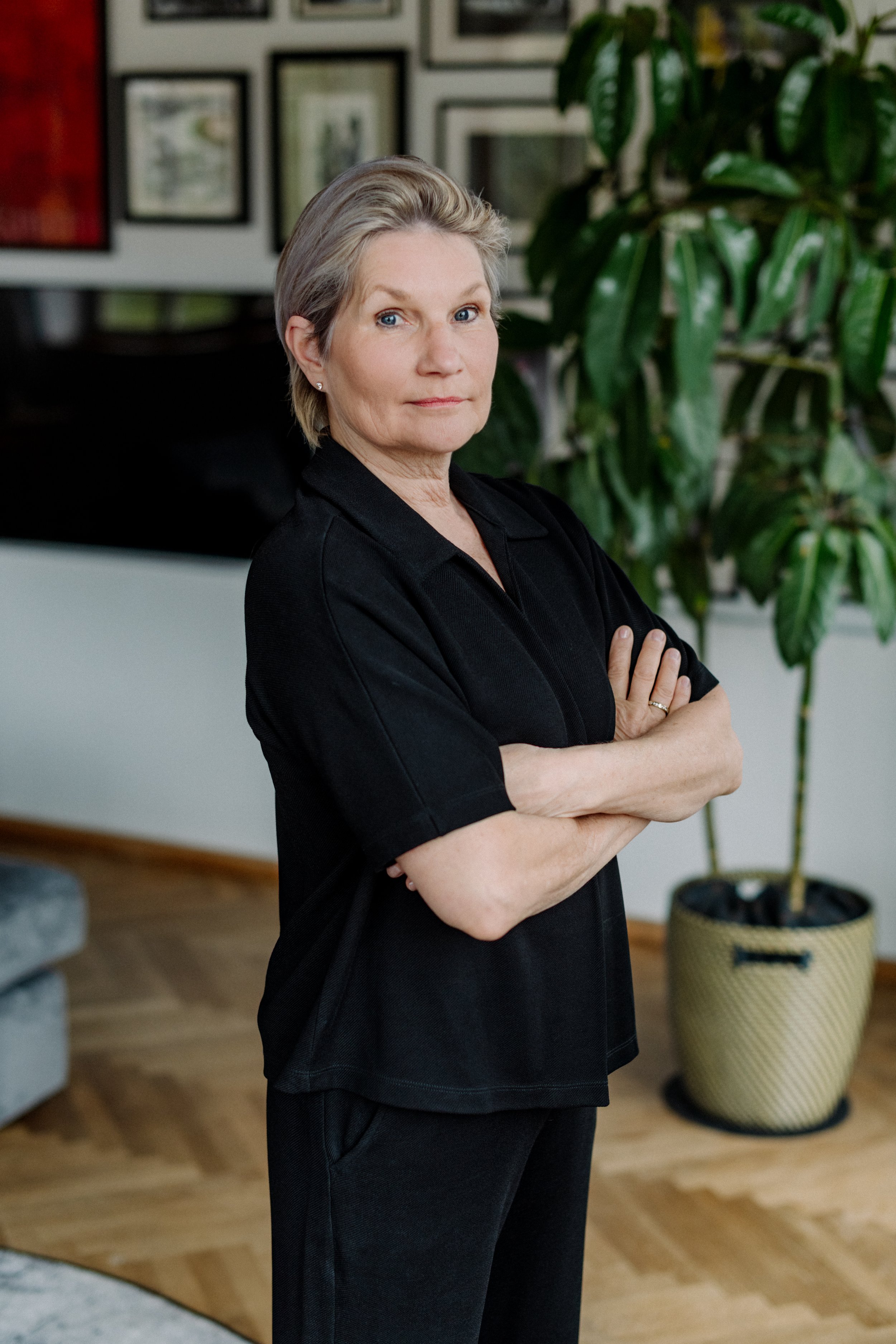Eine ältere Frau mit kurzen, grau-blonden Haaren steht in einem Raum mit einem großen Grünpflanzen im Hintergrund. Sie trägt einen schwarzen Pulli und schwarze Hosen und steht mit verschränkten Armen, Blick direkt in die Kamera.