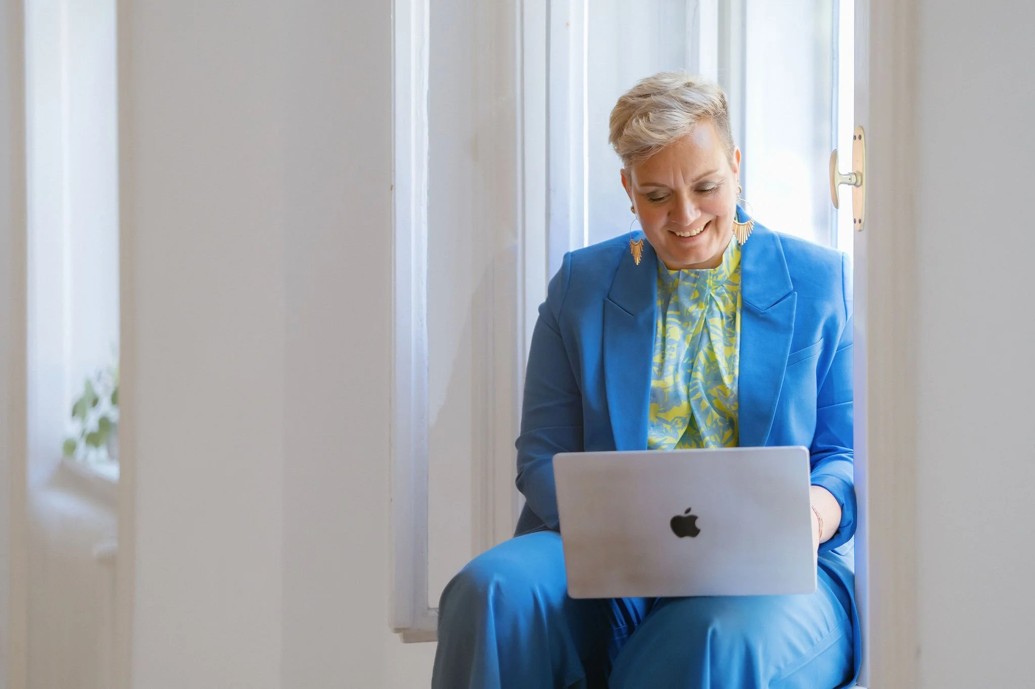 Eine lachende, blond-haarige Frau sitzt auf einer Fensterbank an einem geöffneten Fenster und arbeitet an einem Laptop in einem hellen Raum.