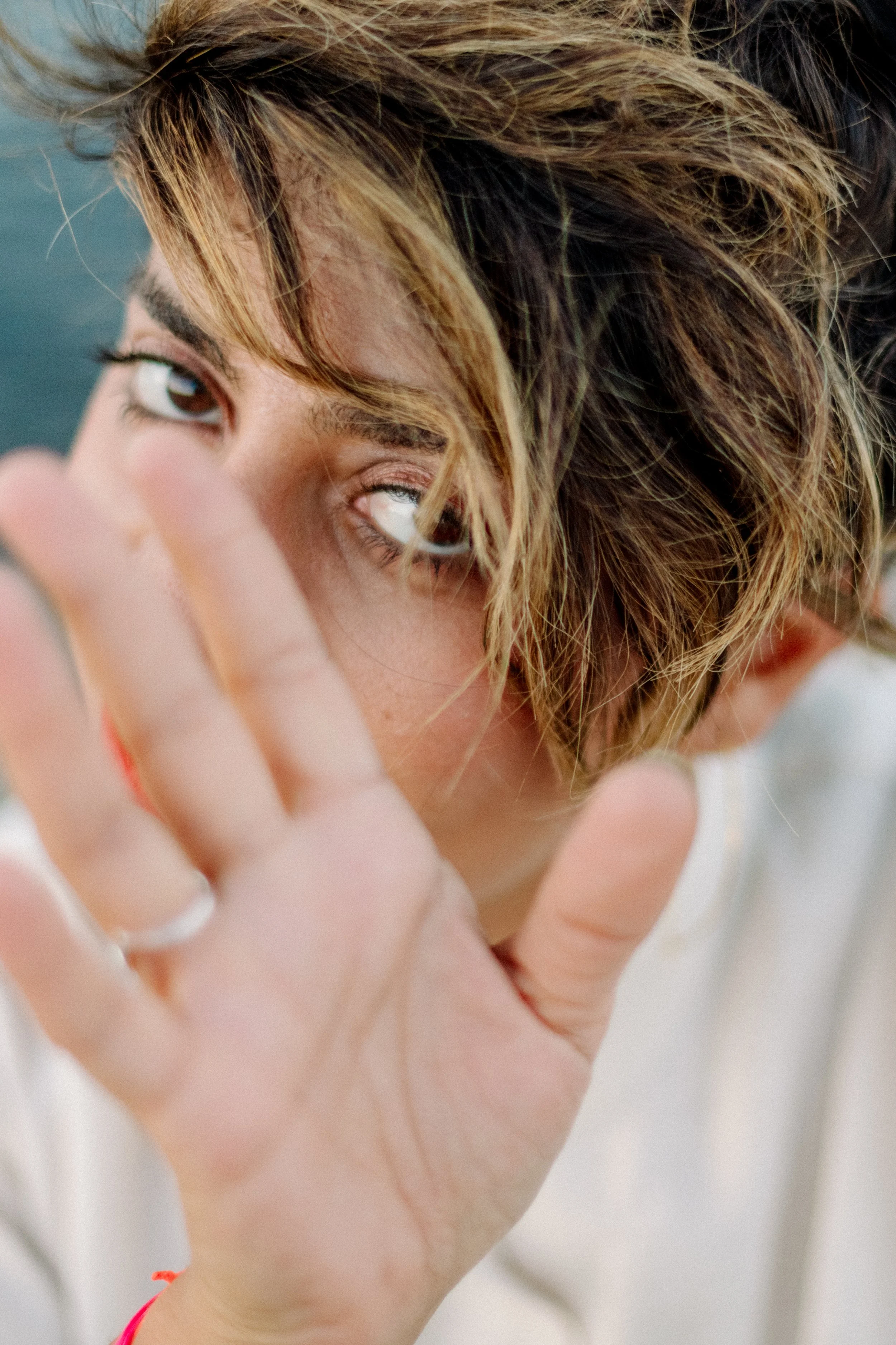 Nahaufnahme einer Person, die eine Hand vor das Gesicht hält. Das Gesicht ist teilweise sichtbar, mit braunen, gewellten Haaren und braunen Augen, die direkt in die Kamera schauen.
