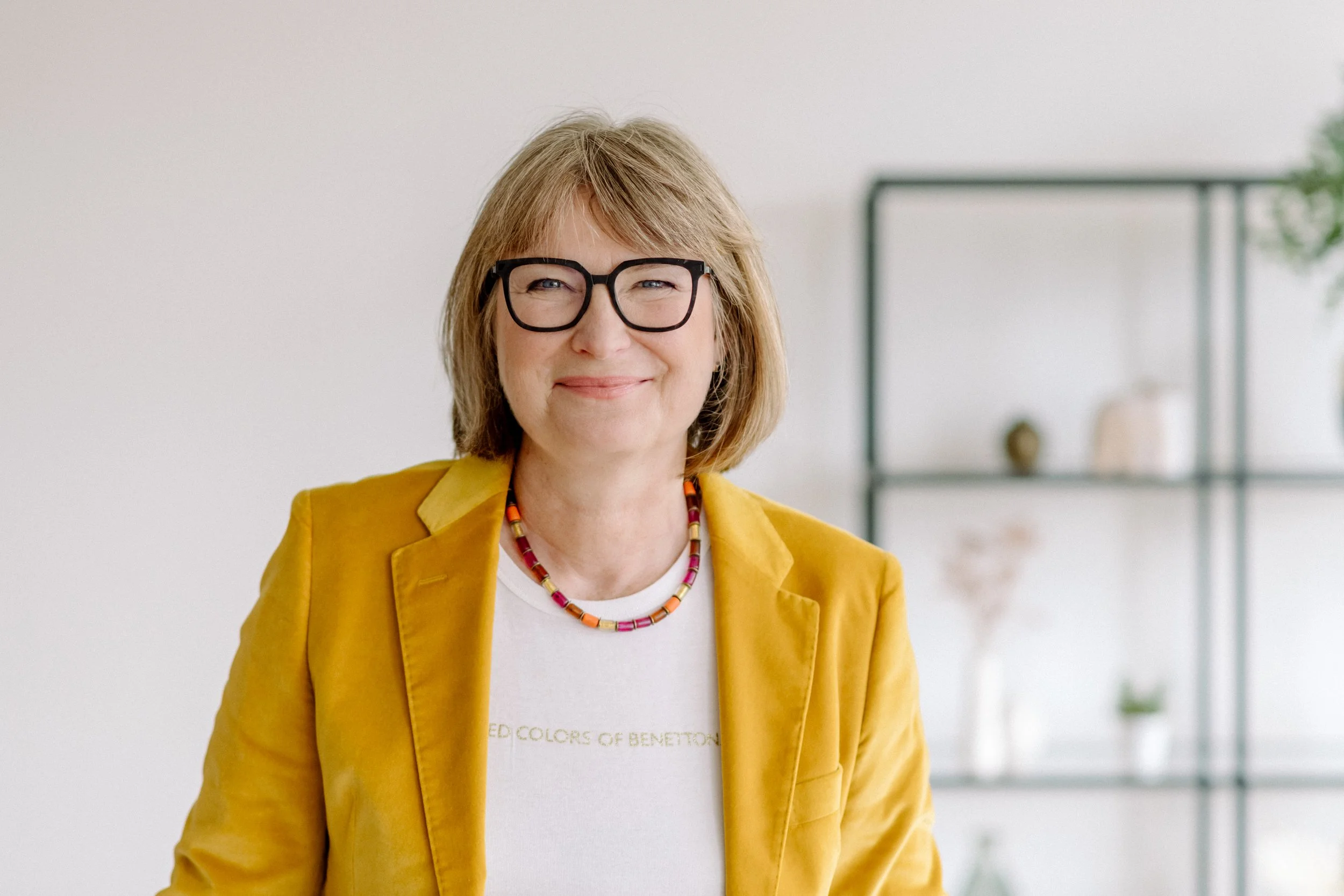 Eine lachende Frau mit kurzem, blonden Haar und schwarzen Brillen, trägt einen gelben Blazer und ein mehrfarbiges Halsband, steht in einem hellen Raum.