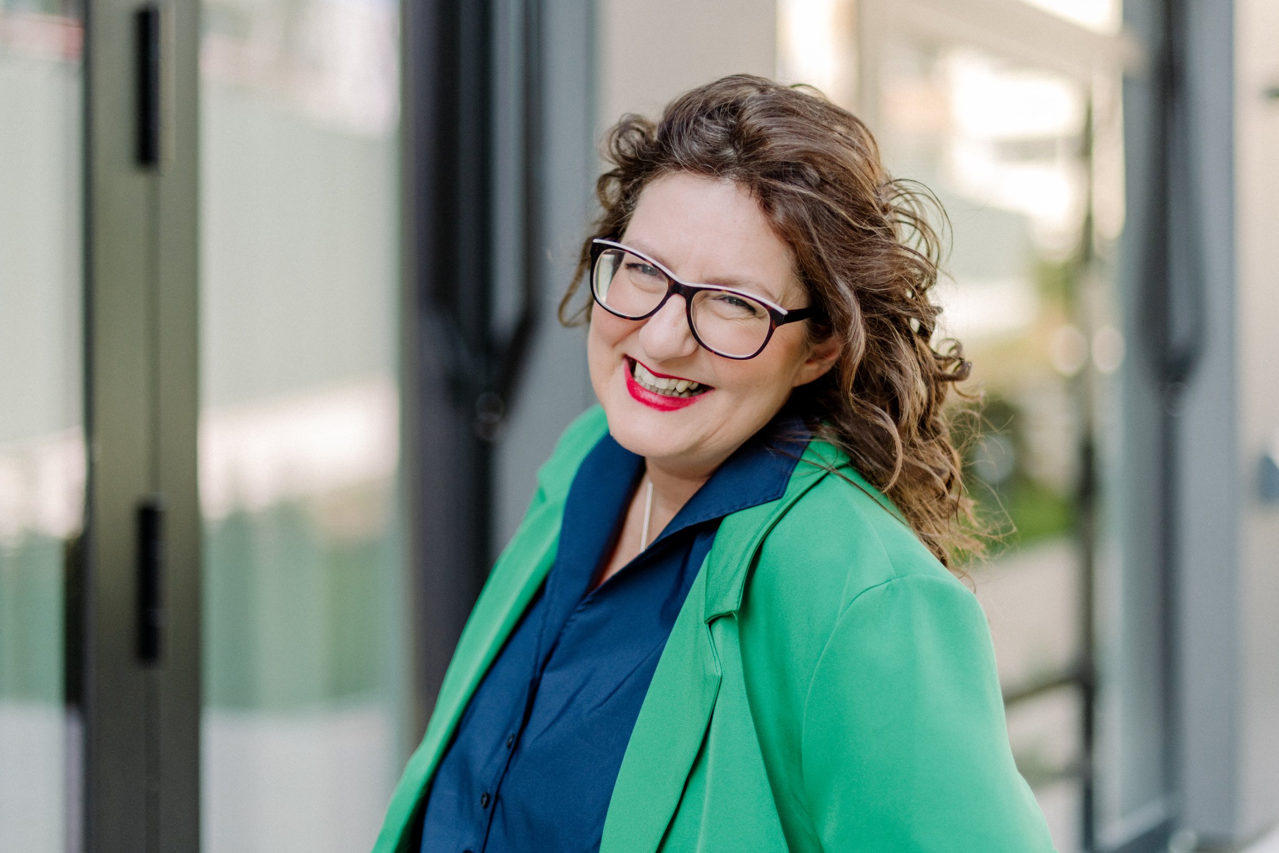 Eine lachende Frau mit Brille, braunen lockigen Haaren, trägt eine grüne Jacke und eine blaue Bluse, steht an einer Bushaltestelle oder einem Bahnhof, im Hintergrund sind Glasfenster sichtbar.