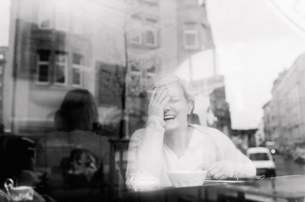 Eine lachende Frau sitzt in einem Café vor einer Fensterscheibe, durch die eine Stadt mit Gebäuden und Autos sichtbar ist.