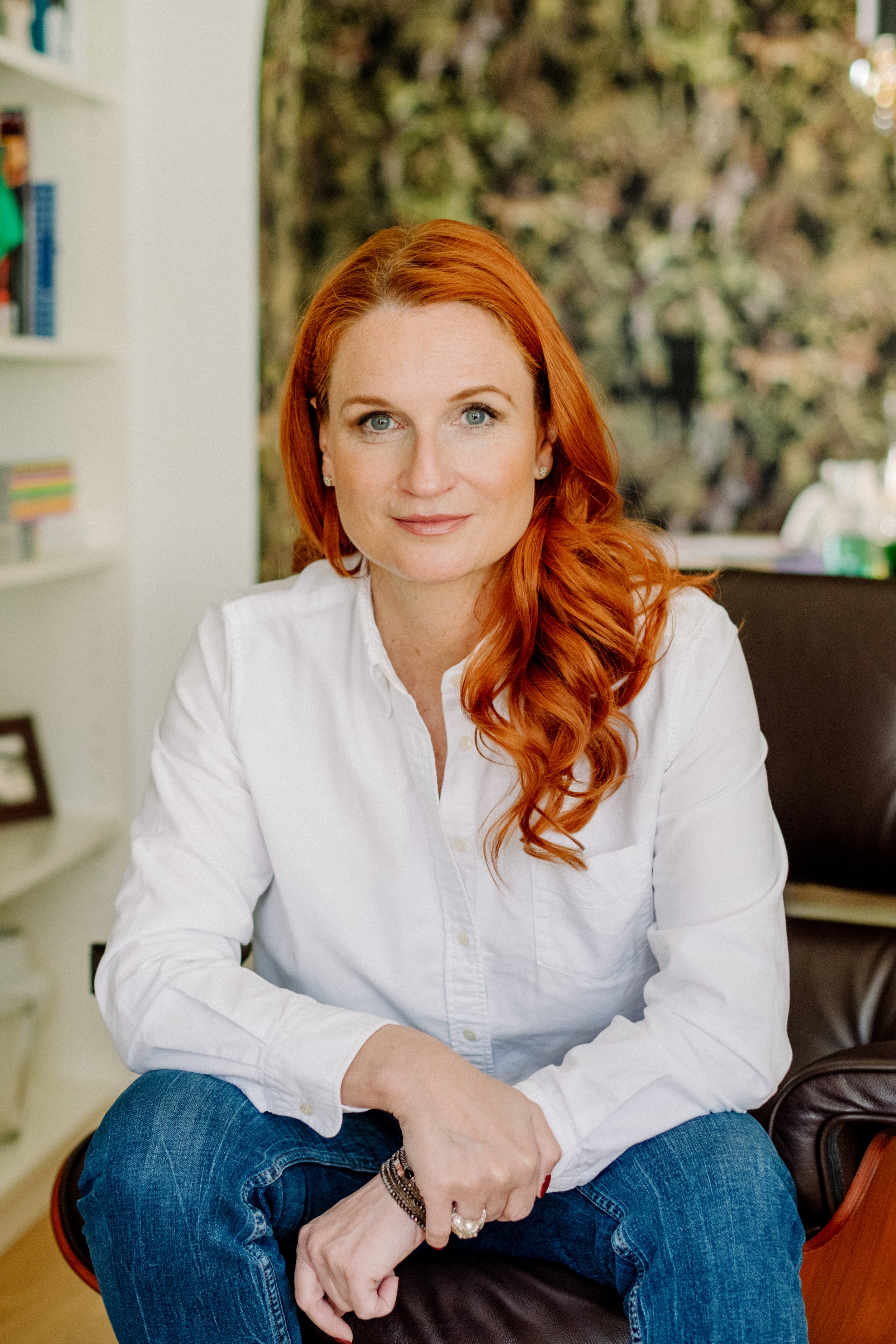 Eine Frau mit roten, gewellten Haaren, trägt eine weiße Bluse und sitzt auf einem Stuhl in einem Raum mit Bücherregal im Hintergrund.