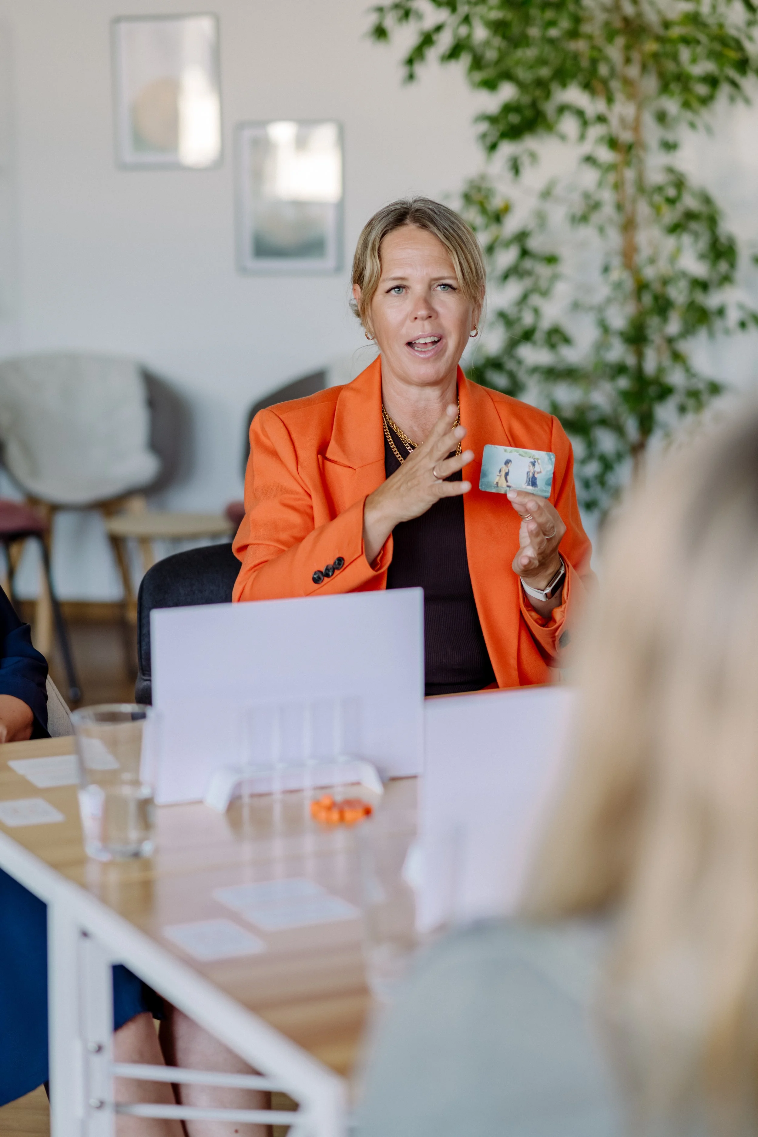Eine Frau in einem orangefarbenen Blazer spricht während eines Meetings, hält eine Spielkarte in der Hand und sitzt an einem Tisch in einem hellen Raum mit Pflanzen und Gemälden an der Wand.