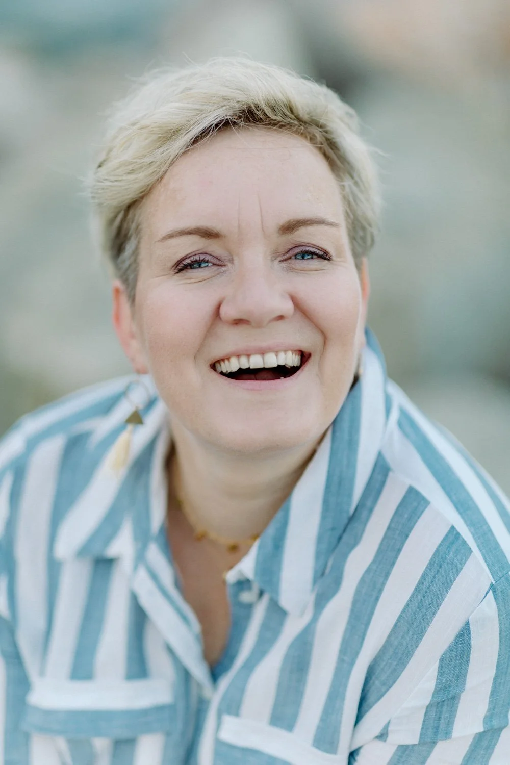 Eine lachende Frau mit kurzen, blondem Haar trägt ein blau-weiß gestreiftes Hemd, die Umgebung ist unscharf, vermutlich im Freien.