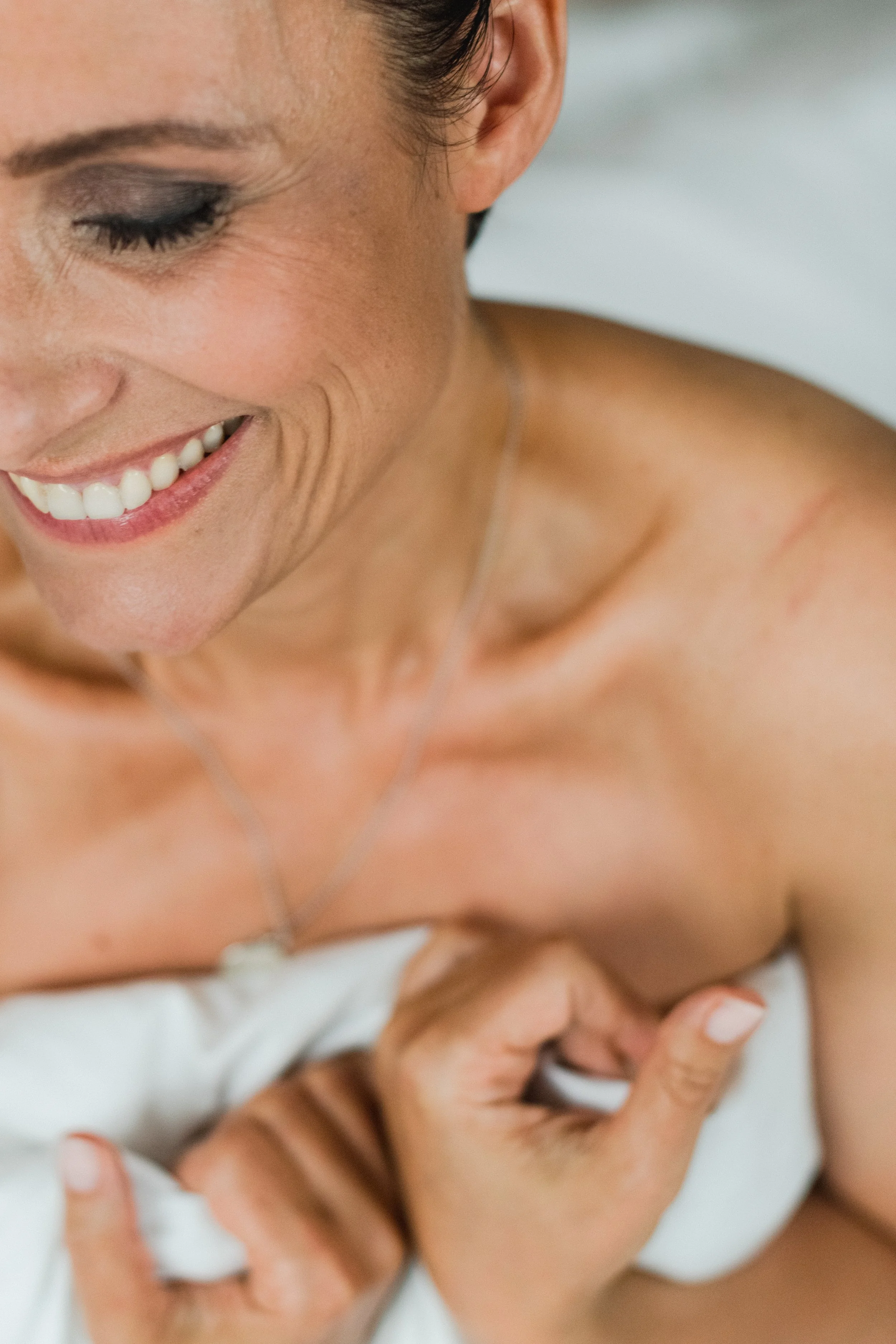 Eine lachende Frau mit dunklem Make-up, die an einem weißen Bett liegt. Das Bild ist eine Nahaufnahme ihres Gesichts, ihrer Schulter und ihrer Hand. Der Fokus liegt auf ihrem Gesicht und Lächeln.