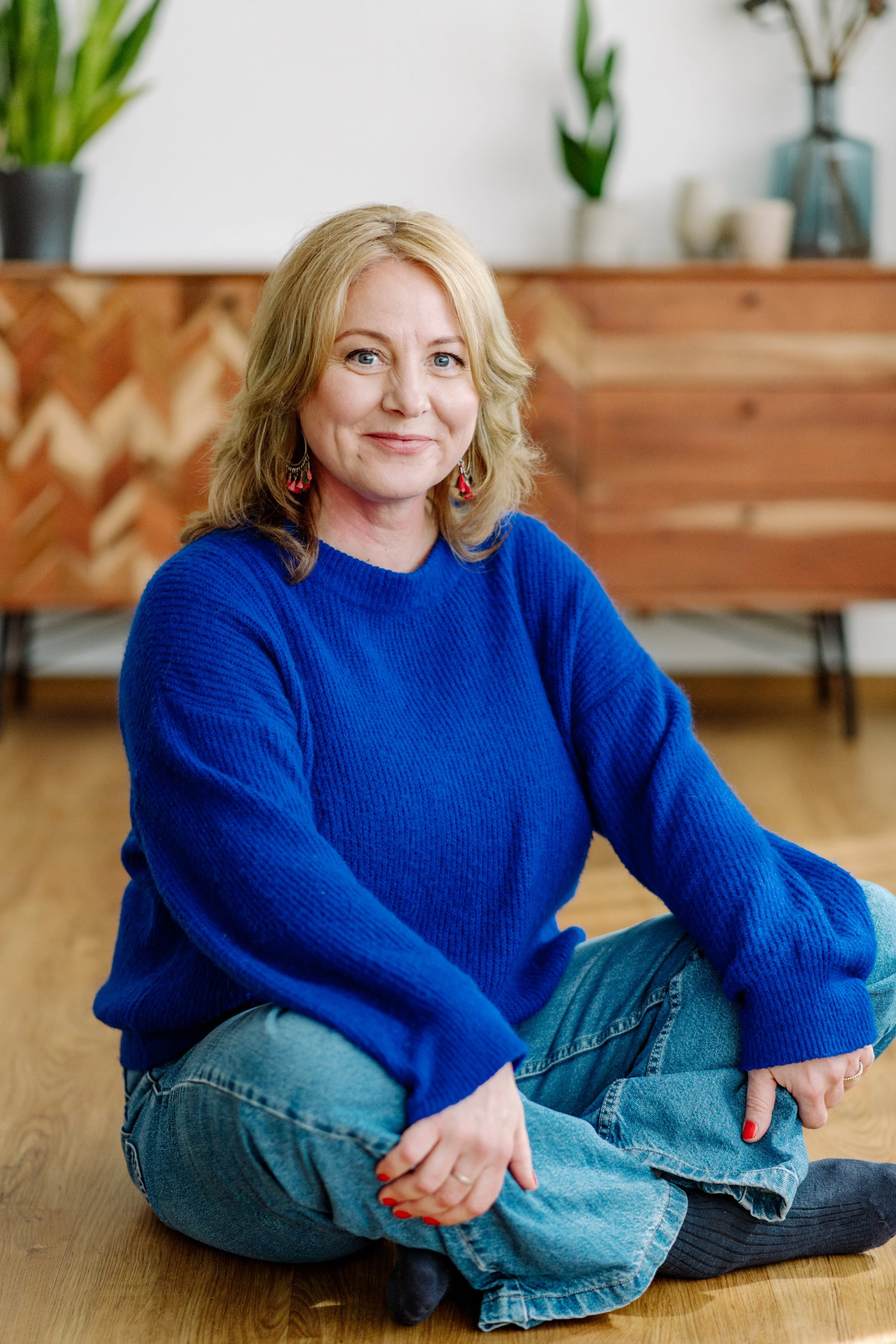 Eine Frau sitzt im Casual-Look auf einem Holzboden, trägt eine blaue Strickjacke, Jeans und schwarze Socken, im Hintergrund sind Pflanzen und Vasen sichtbar.